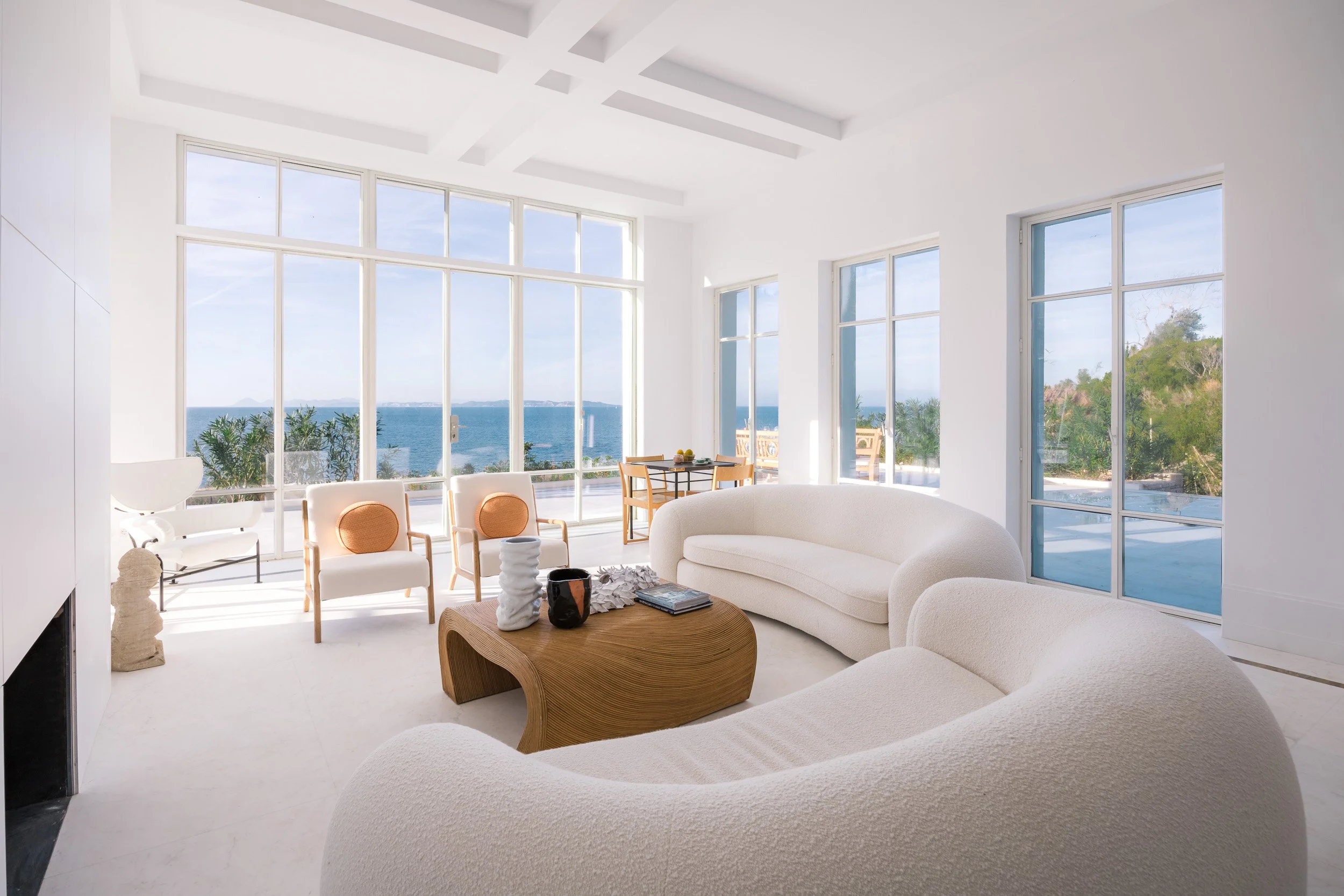 Bright living room with large windows showcasing a view of the ocean, featuring white curved couches, wooden coffee table, armchairs with orange cushions, and a small table with fruit in the background.