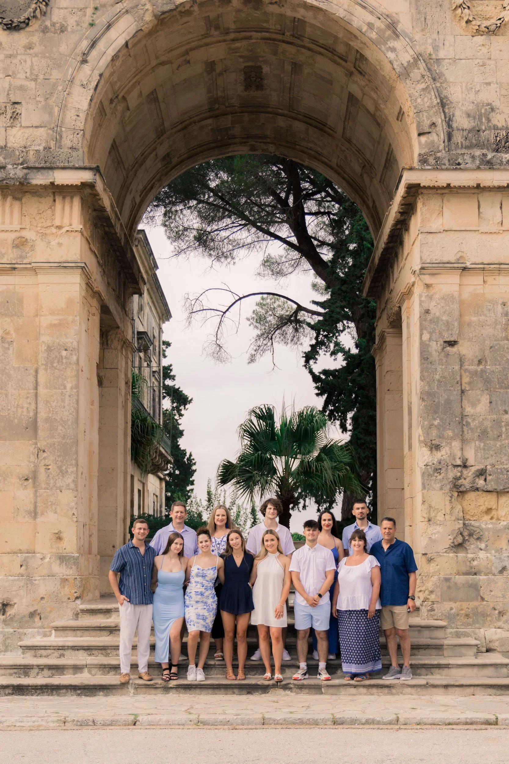 A group of fourteen people standing on stone steps under an ancient stone arch, with trees in the background, during daytime.