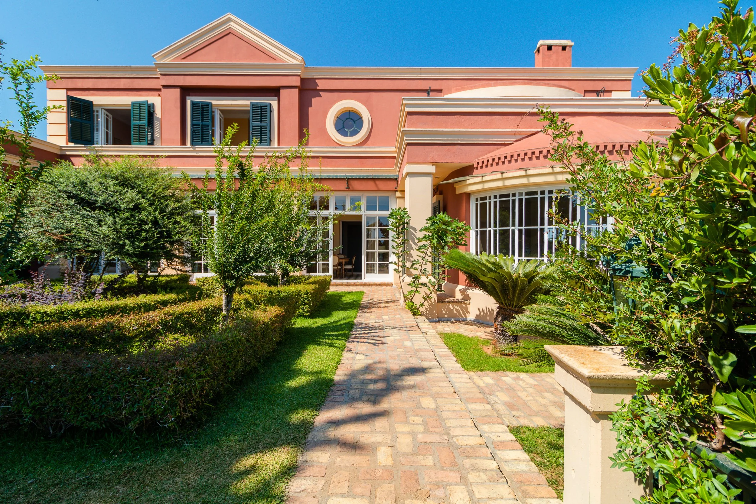 Front view of a pink house with a brick pathway leading to the entrance, lush green shrubs, and trees, under a clear blue sky.