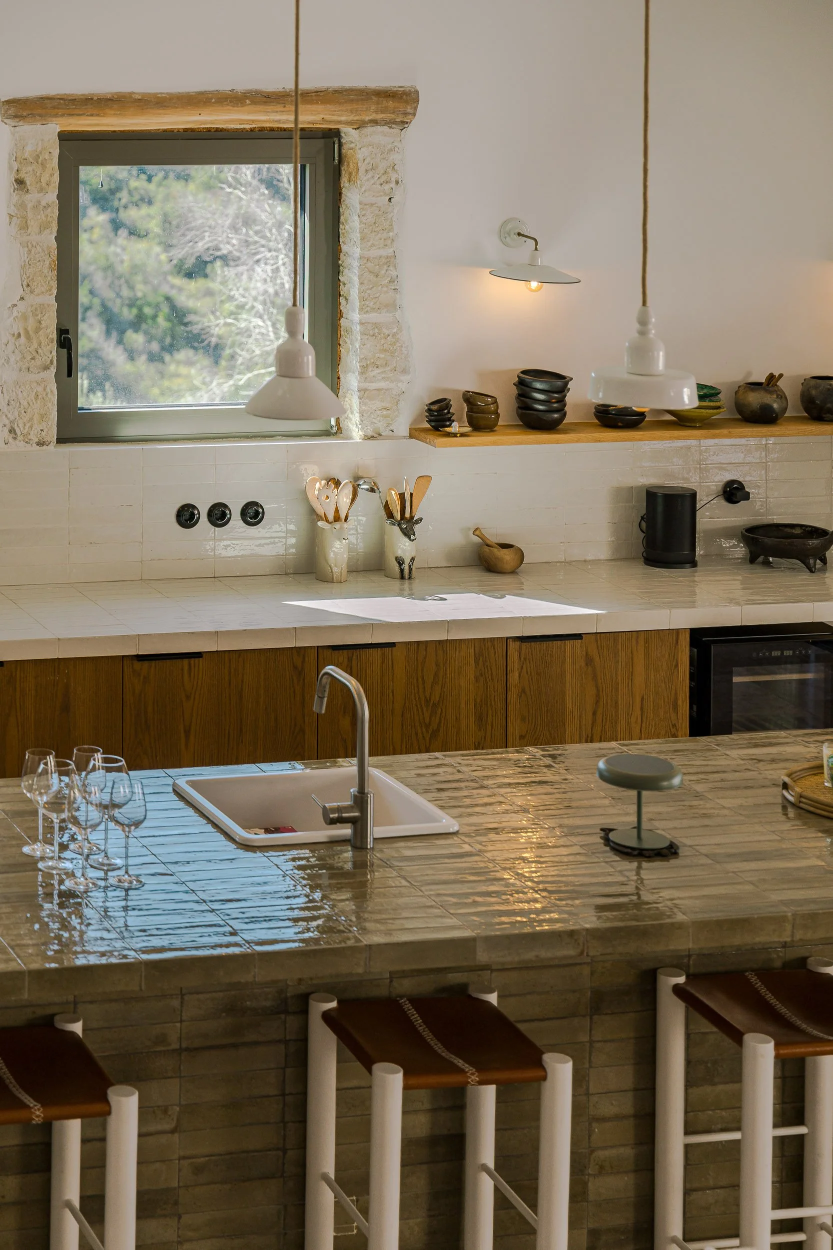 Modern kitchen with a large tiled island, wooden stools, a sink, wine glasses, and various bowls and kitchenware displays near a window with stone framing.