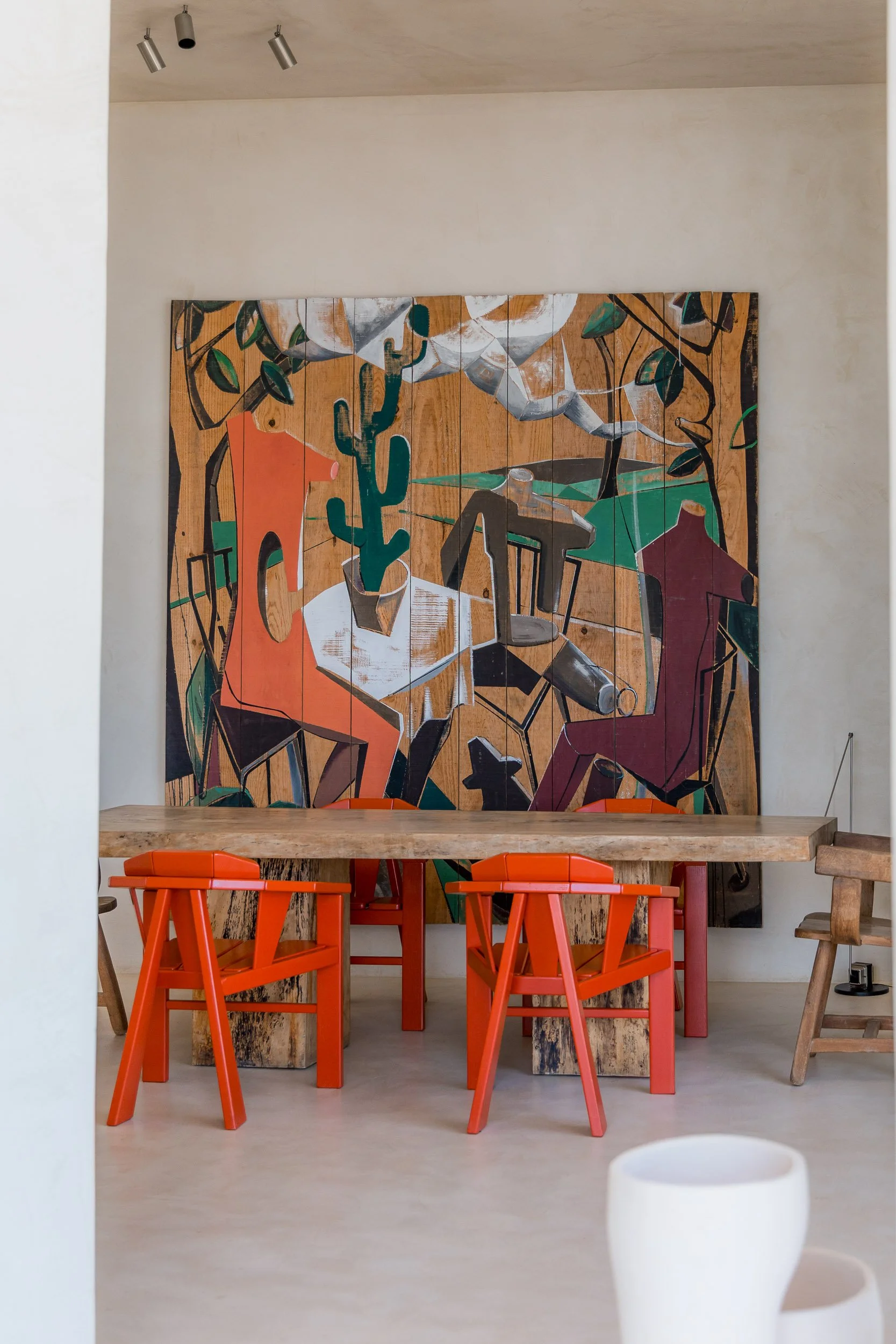 Interior dining space with a large abstract painting hanging on the wall, a rustic wooden table, and orange chairs in the foreground.