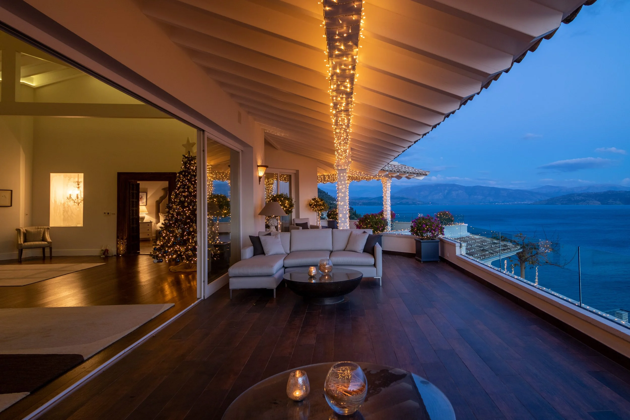 A spacious balcony decorated for Christmas with string lights hanging from the ceiling, a white sectional sofa with pillows, potted plants, and a view of the ocean and mountains at dusk.