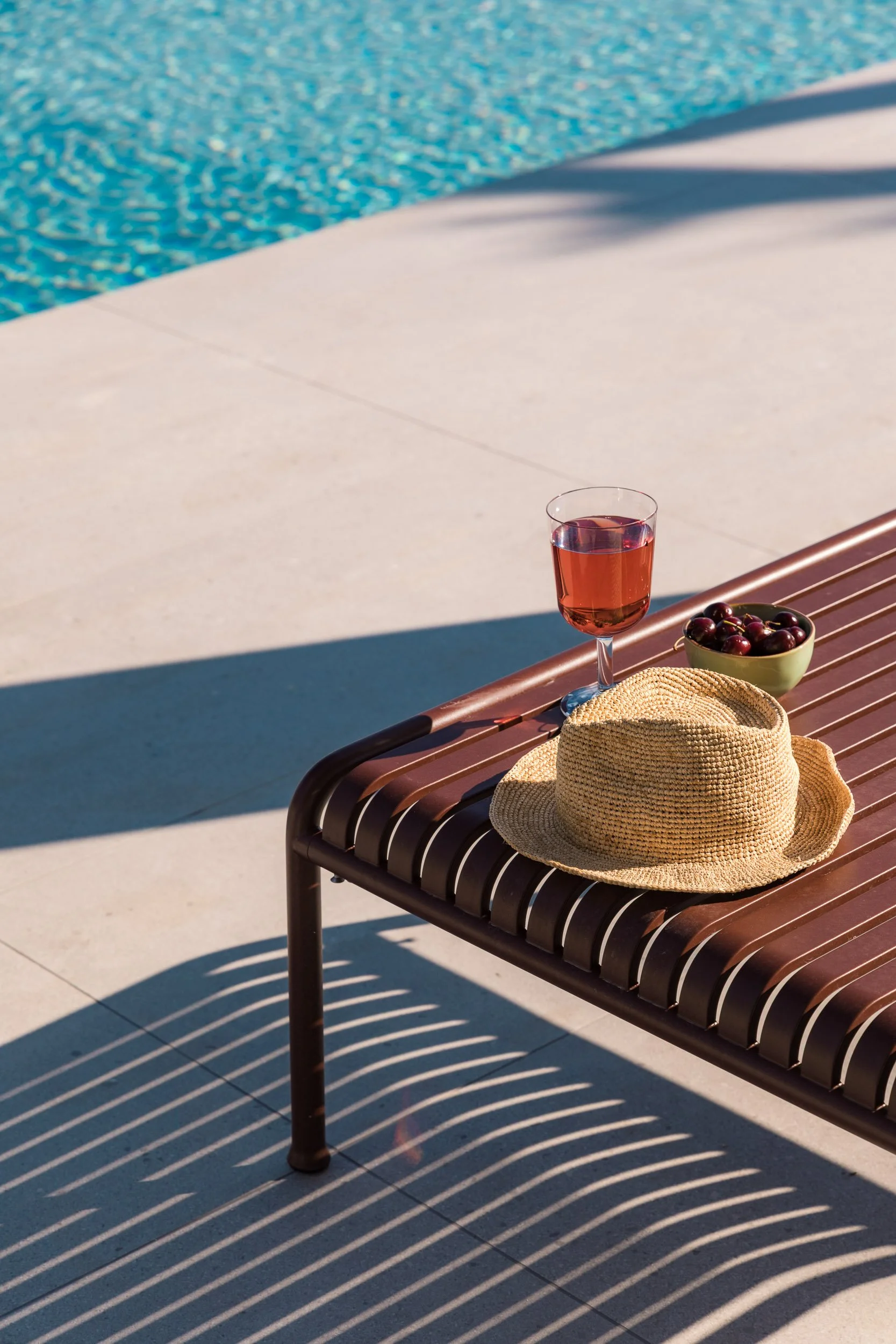 Poolside scene with a brown slatted lounge chair holding a straw hat, a glass of pink drink, a bowl of cherries, and a pool in the background.