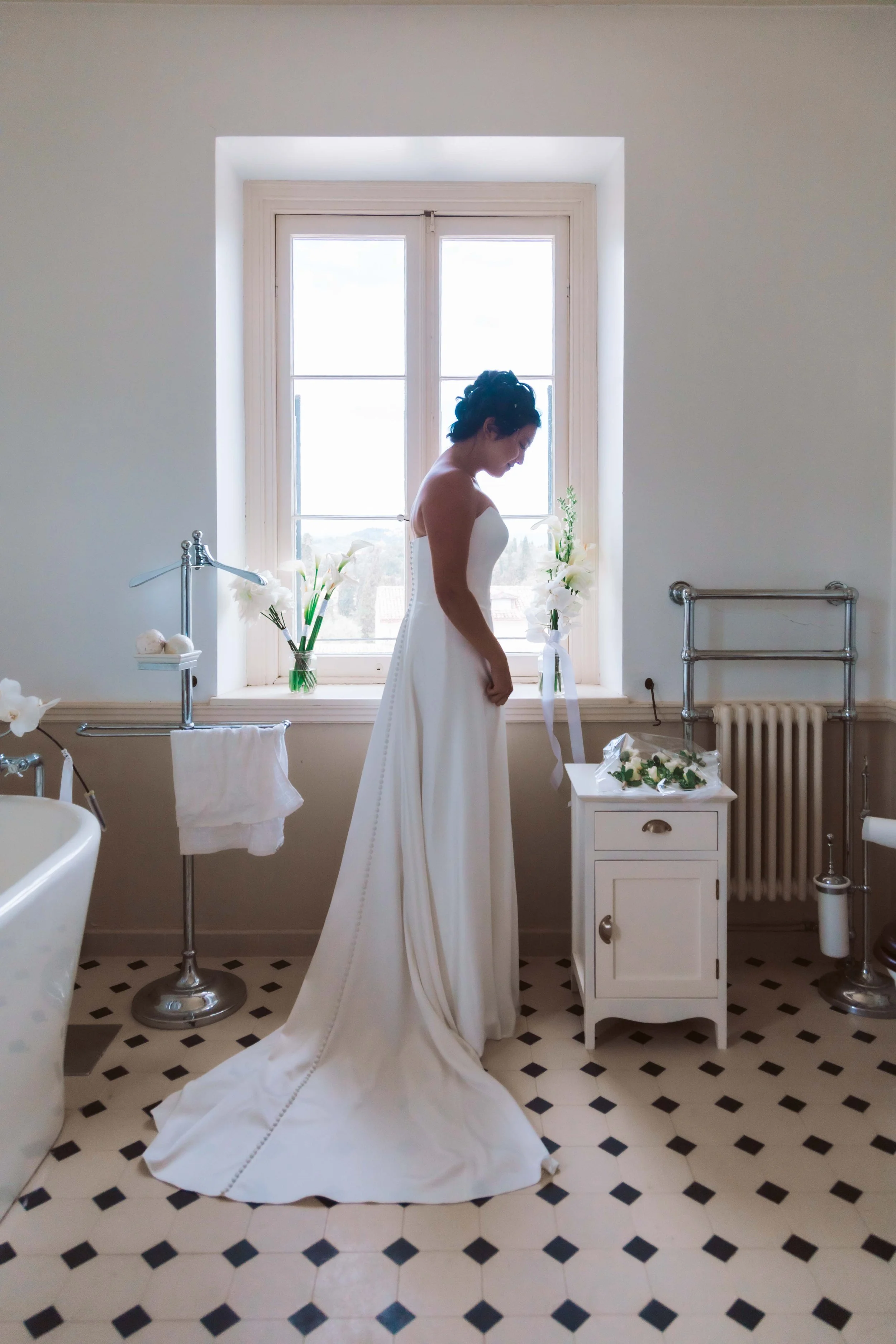 A bride in a white wedding dress standing in a bathroom by a window, with flowers and a small cabinet nearby.
