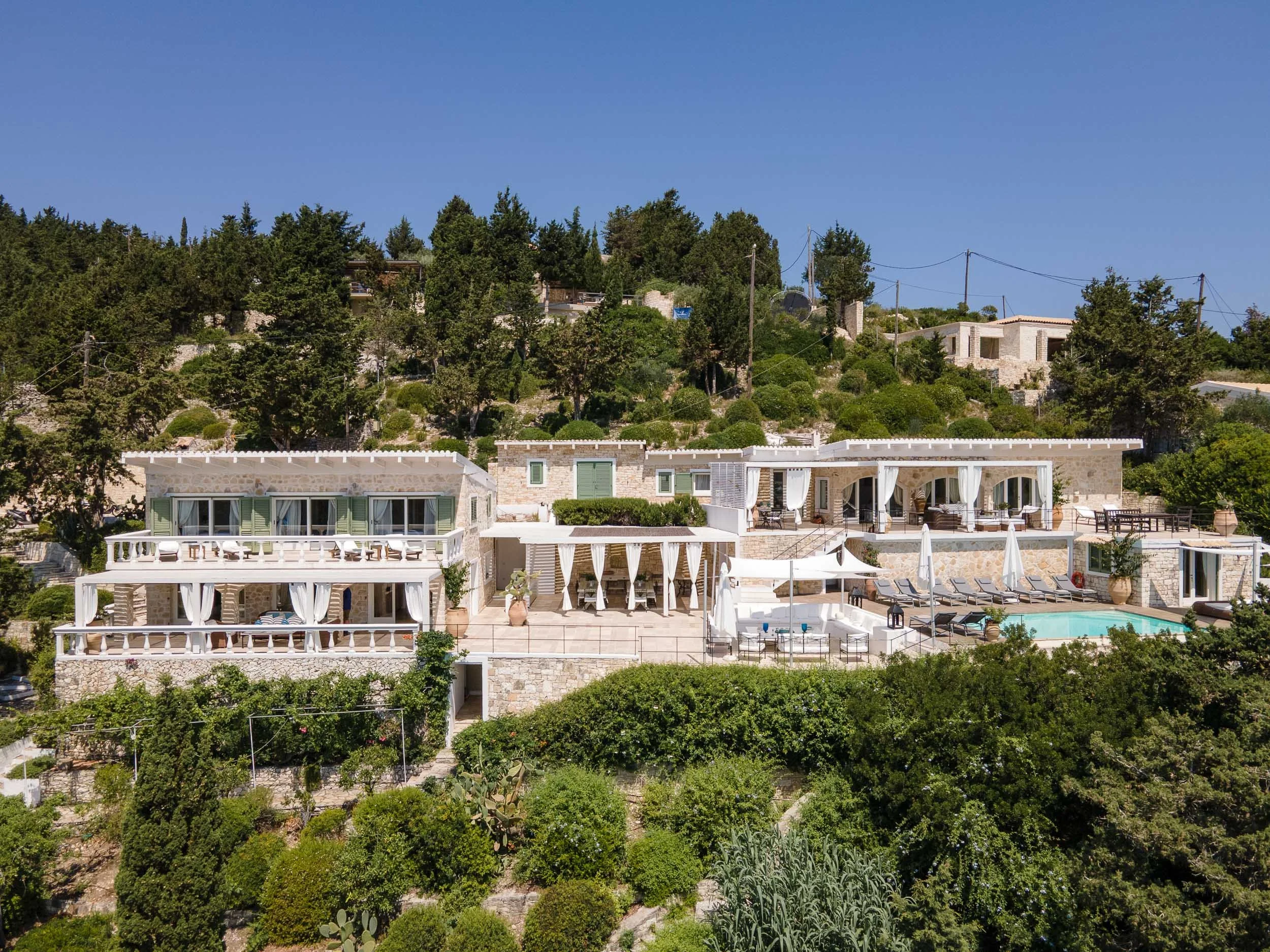A hillside hotel with multiple levels of white buildings surrounded by lush greenery, featuring outdoor terraces, cabanas, and a swimming pool with lounge chairs.