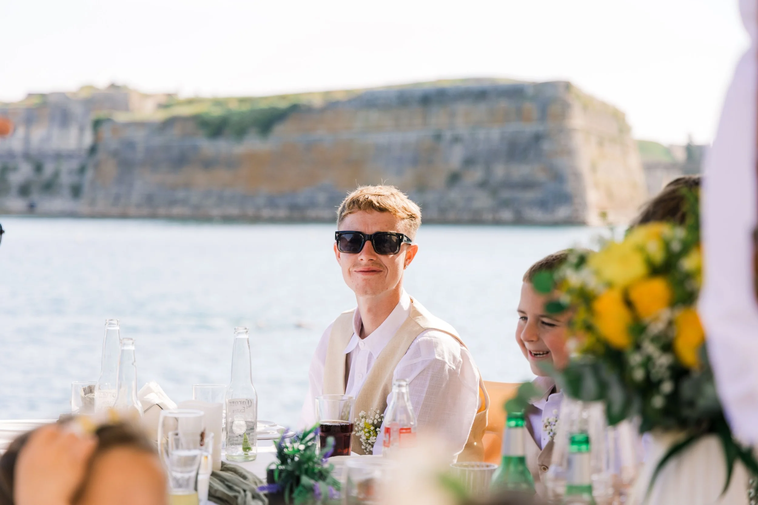People celebrating at an outdoor event near the water with a large fortress in the background, including a man wearing sunglasses and a child smiling.