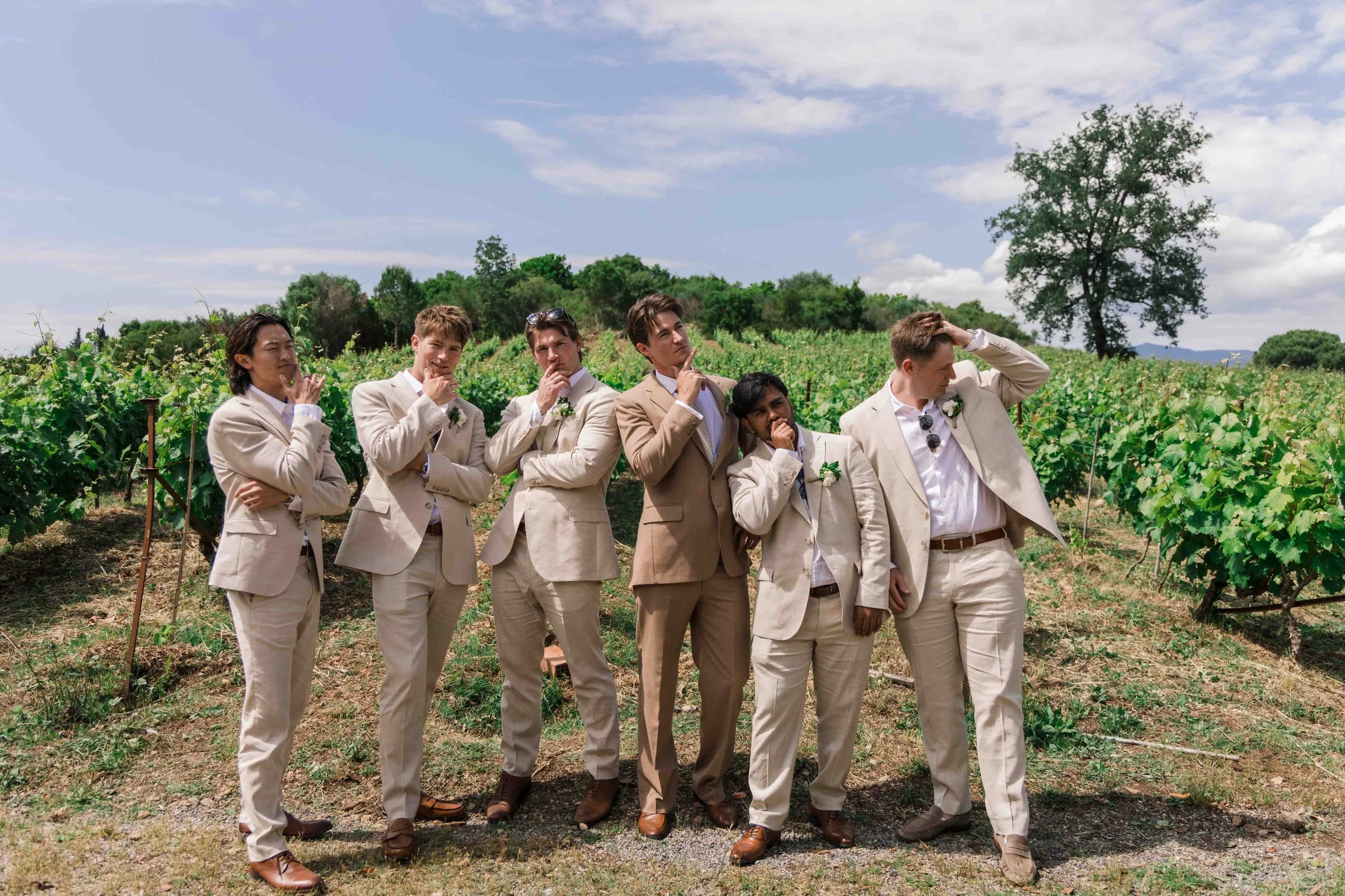 A group of seven men in beige suits standing in a vineyard on a sunny day, posing with thoughtful and playful expressions.