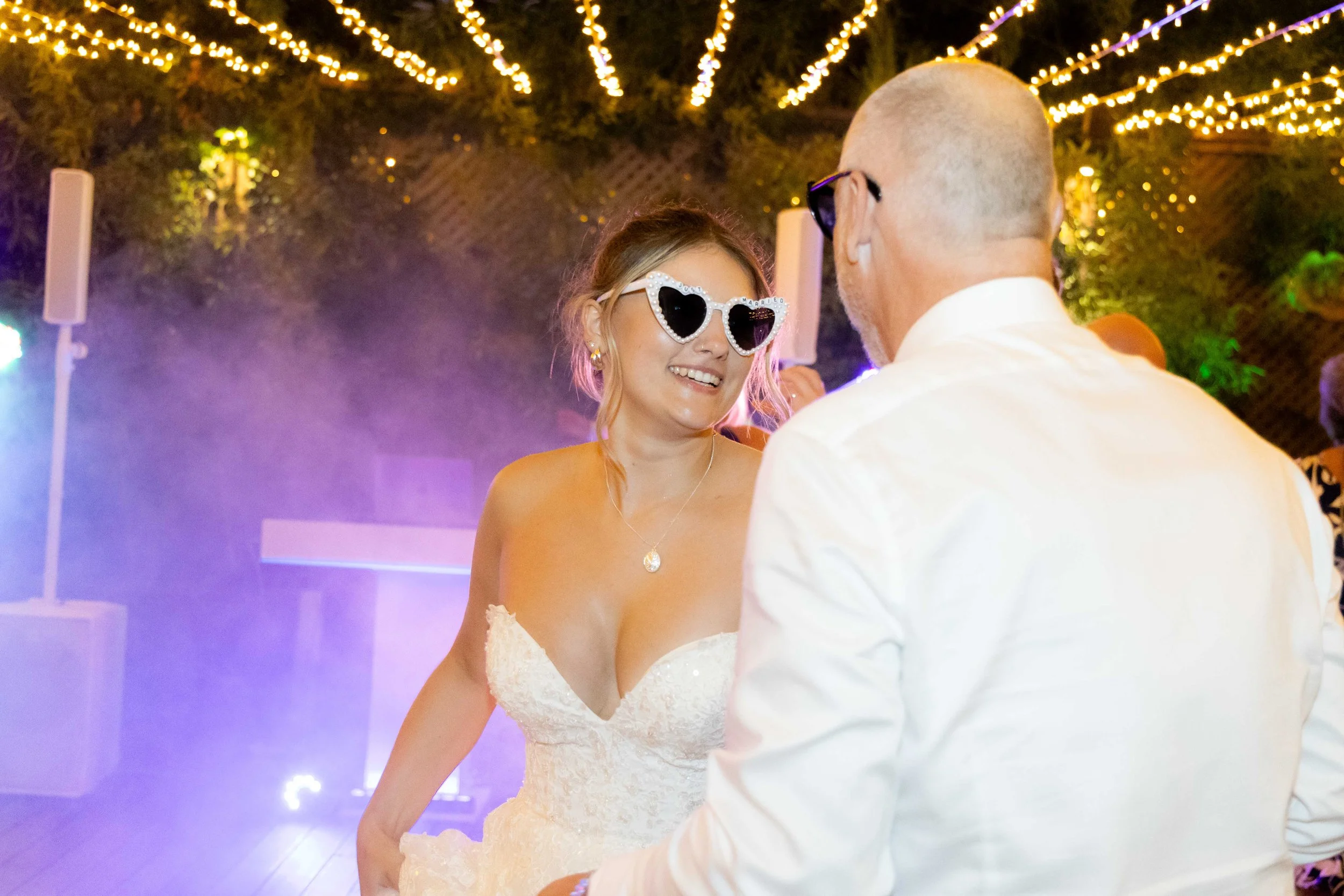 A bride wearing heart-shaped sunglasses and a wedding dress smiling at a man in a white shirt at a wedding reception with string lights and purple light effects in the background.