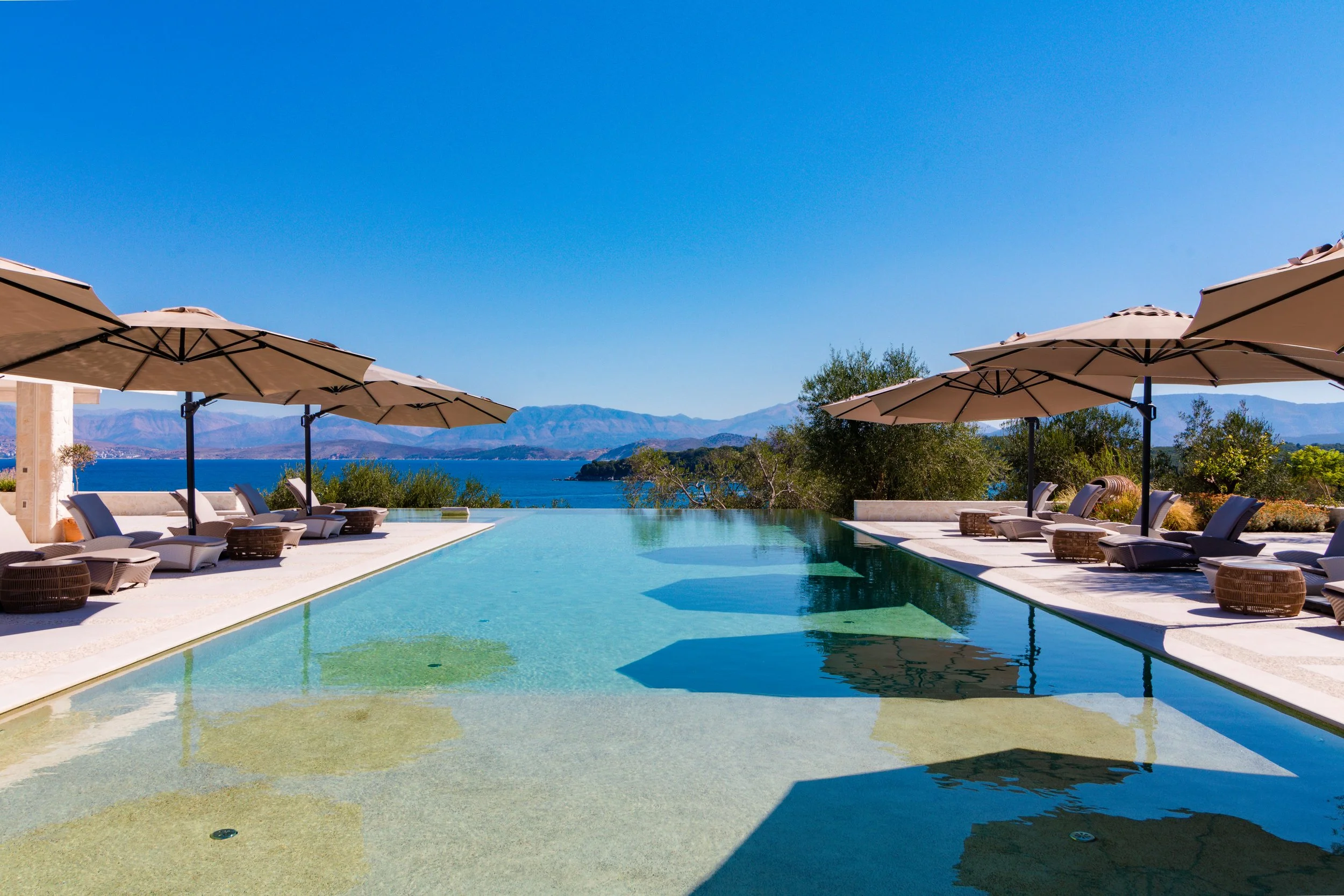 Infinity pool with lounge chairs and umbrellas overlooking a mountain and lake view under a clear blue sky.