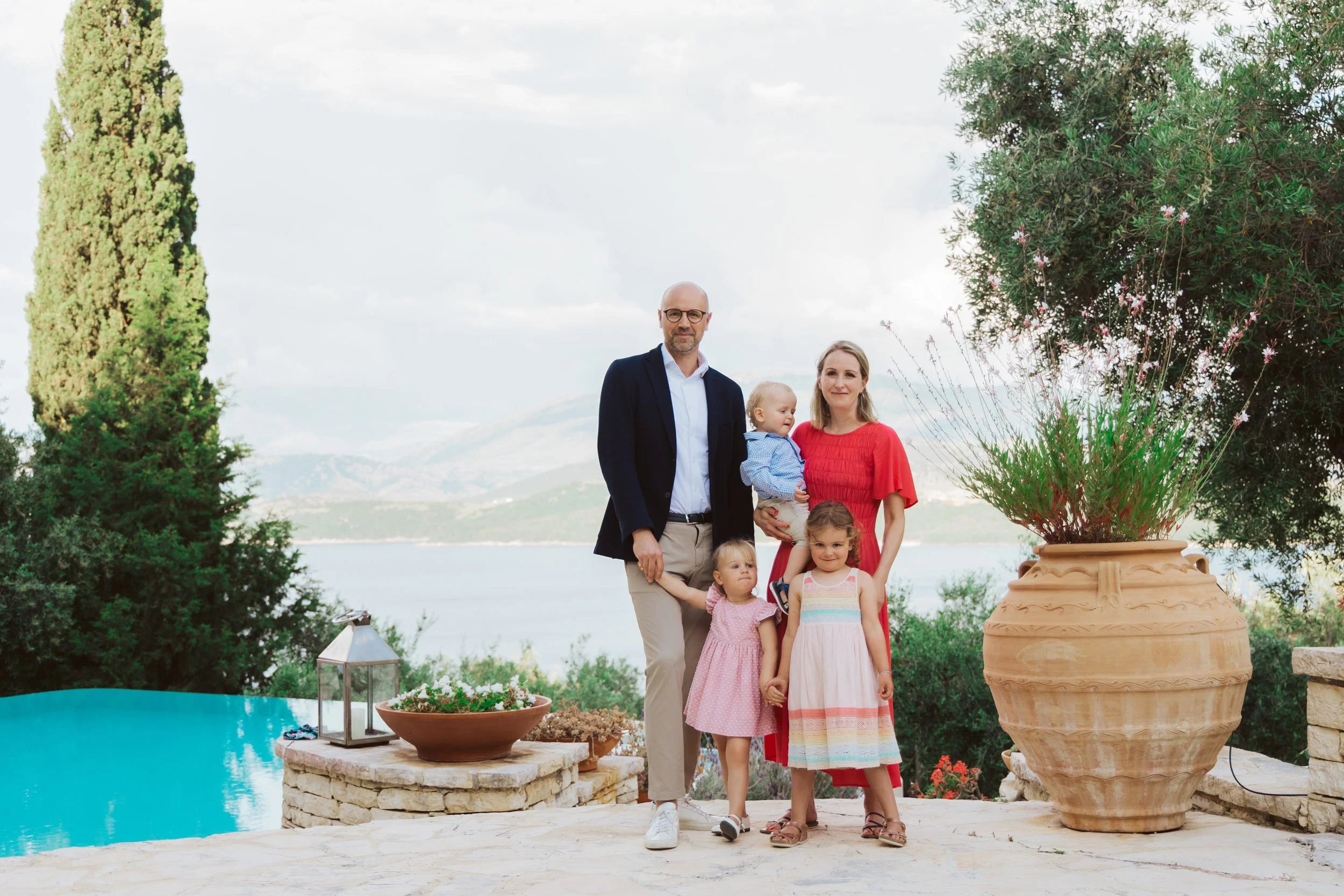 A family of five stands outdoors by a pool with mountains in the background. A man, woman, and three young girls pose for a photo.