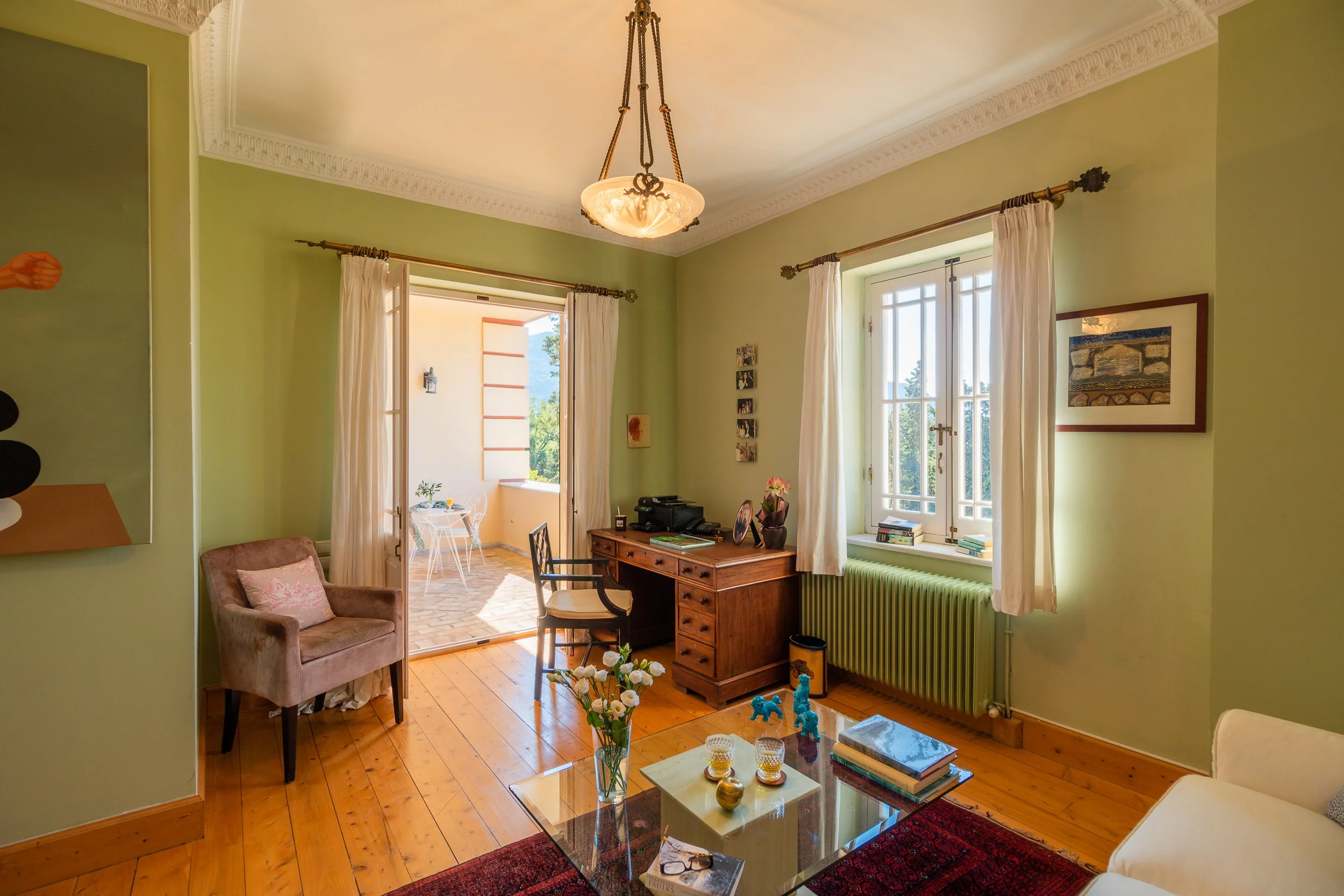 Living room with green walls, wooden floor, white curtains, windows, a wooden desk, a beige armchair with pink pillow, a glass coffee table with books and decorative items, and a small balcony with outdoor furniture.