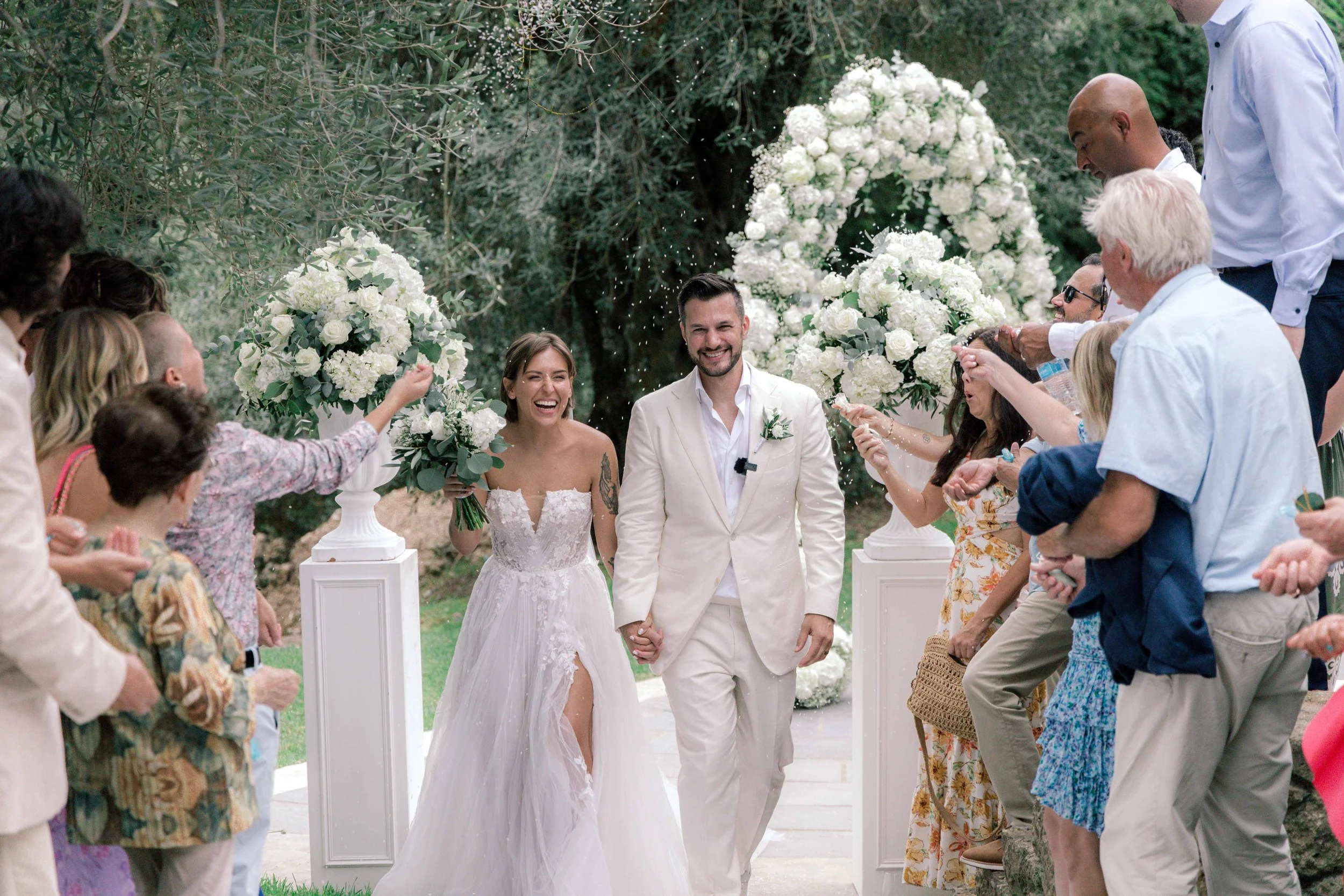 A newly married couple walking down the aisle, holding hands, surrounded by friends and family celebrating with flower confetti outdoors.