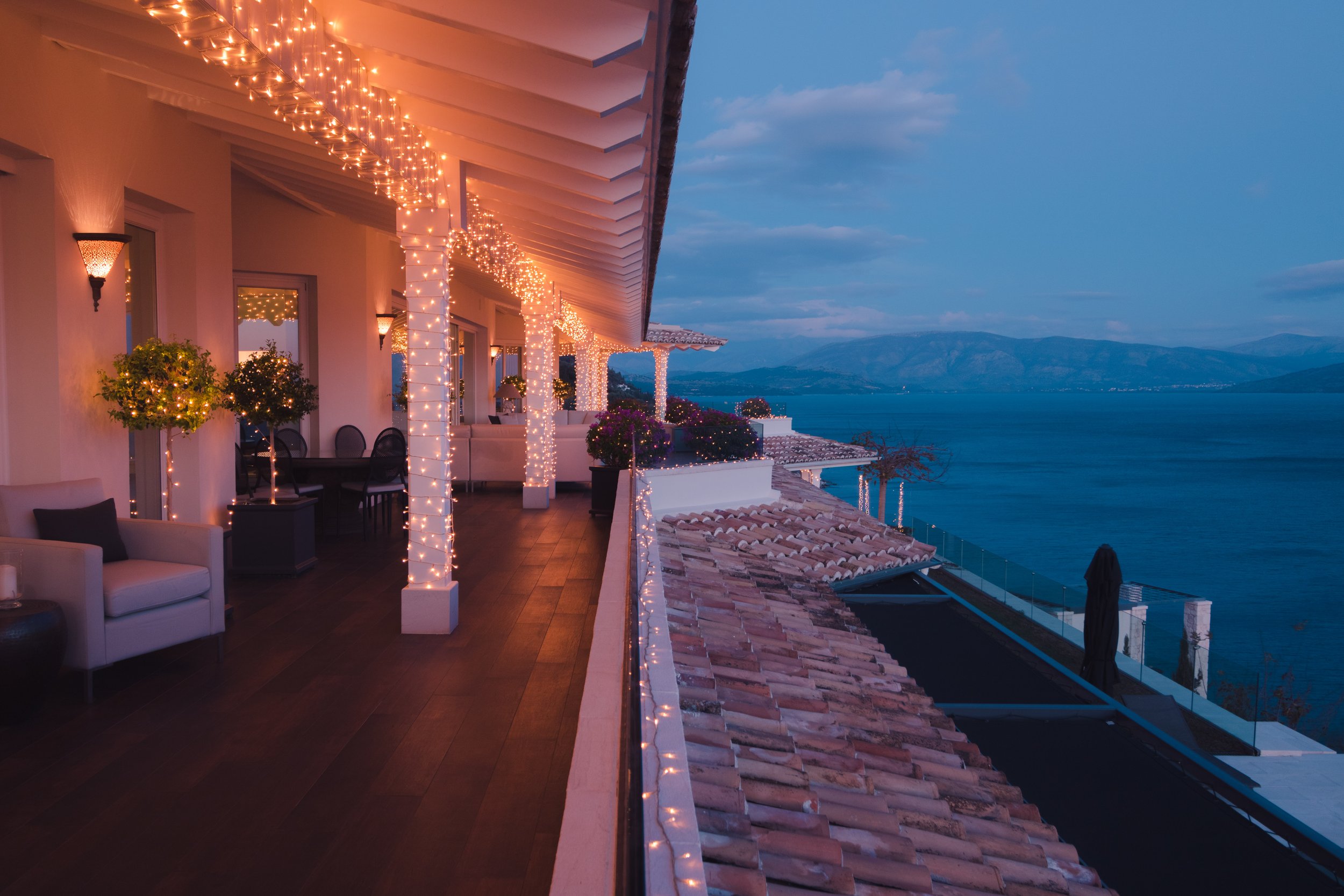 A spacious outdoor balcony decorated with string lights wrapped around white pillars, overlooking a body of water with mountains in the distance during dusk. The balcony featurespotted plants and outdoor furniture, including white couches and a table