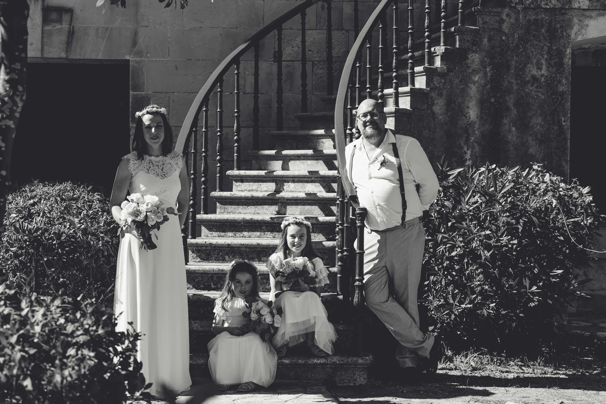 A wedding couple and two young girls with floral bouquets standing on a stone staircase outside, with a wrought iron railing and bushes nearby.