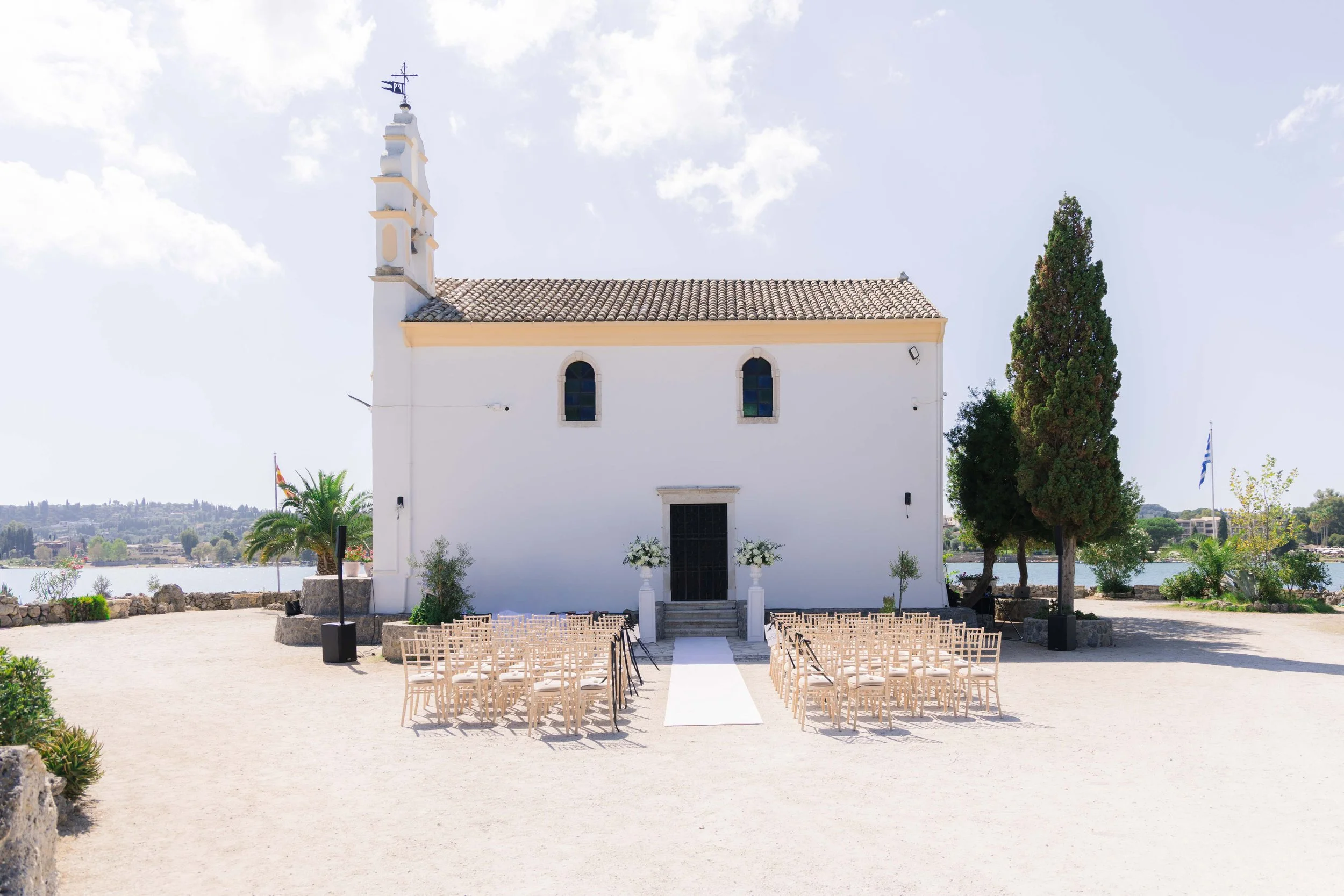 White chapel with two stained glass windows, decorated for a wedding with chairs arranged outside, floral arrangements, and a white aisle runner leading to the entrance, near a body of water.