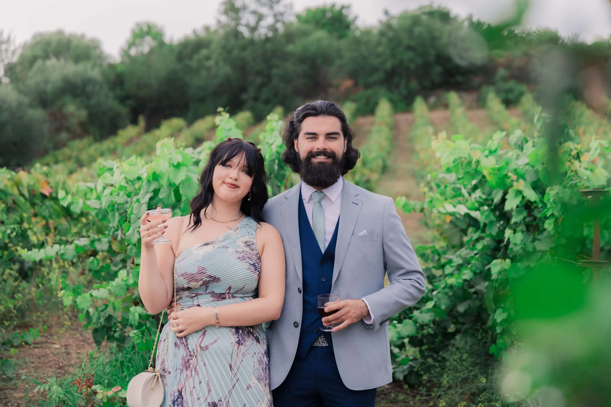 A man and woman standing together in a vineyard, both holding drinks and smiling at the camera during a sunny day.