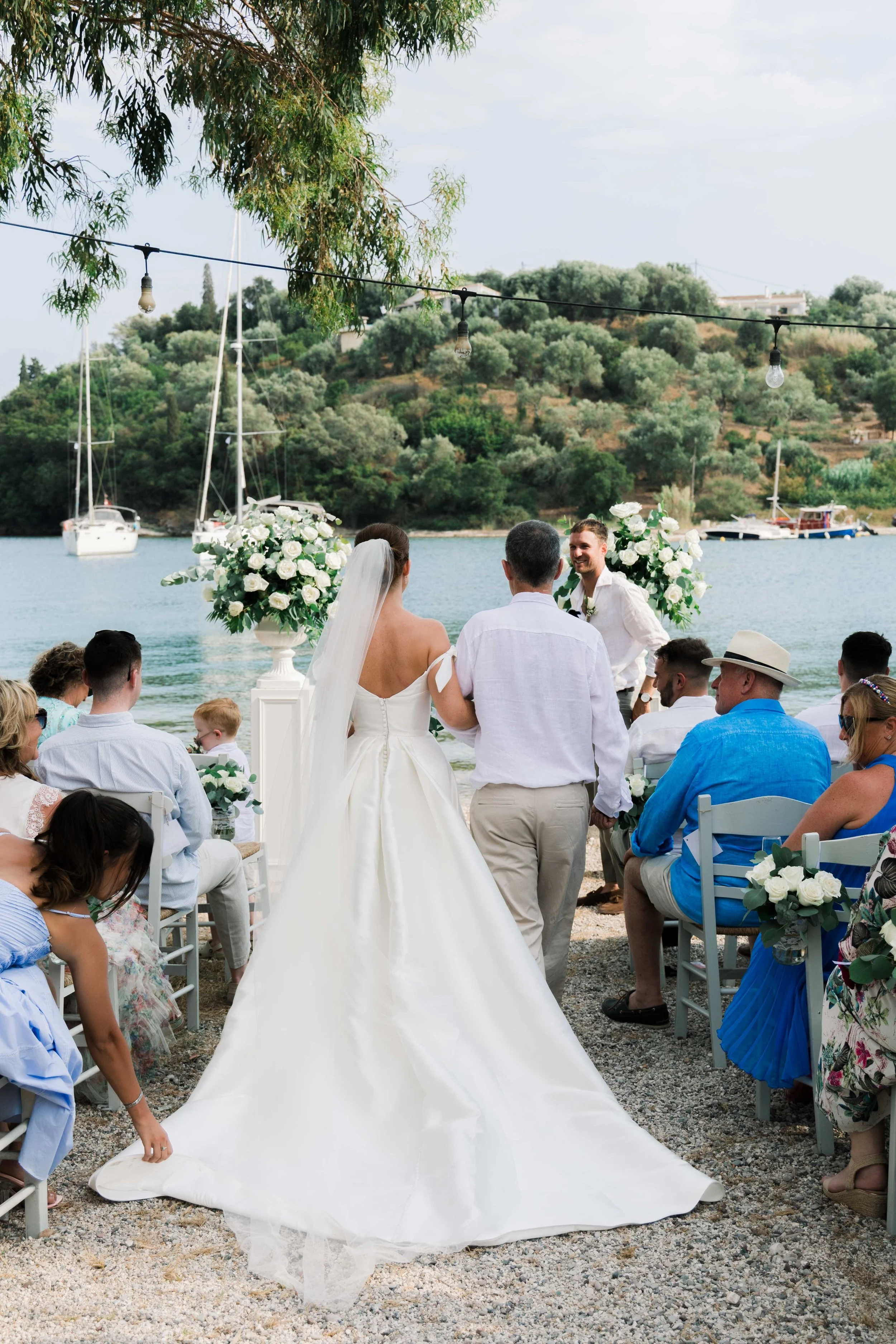 A wedding ceremony taking place by the water with sailboats and boats docked in the background. The bride in a white gown and veil walks down the aisle with a man, over a gravel path lined with guests seated on both sides. The ceremony is decorated w