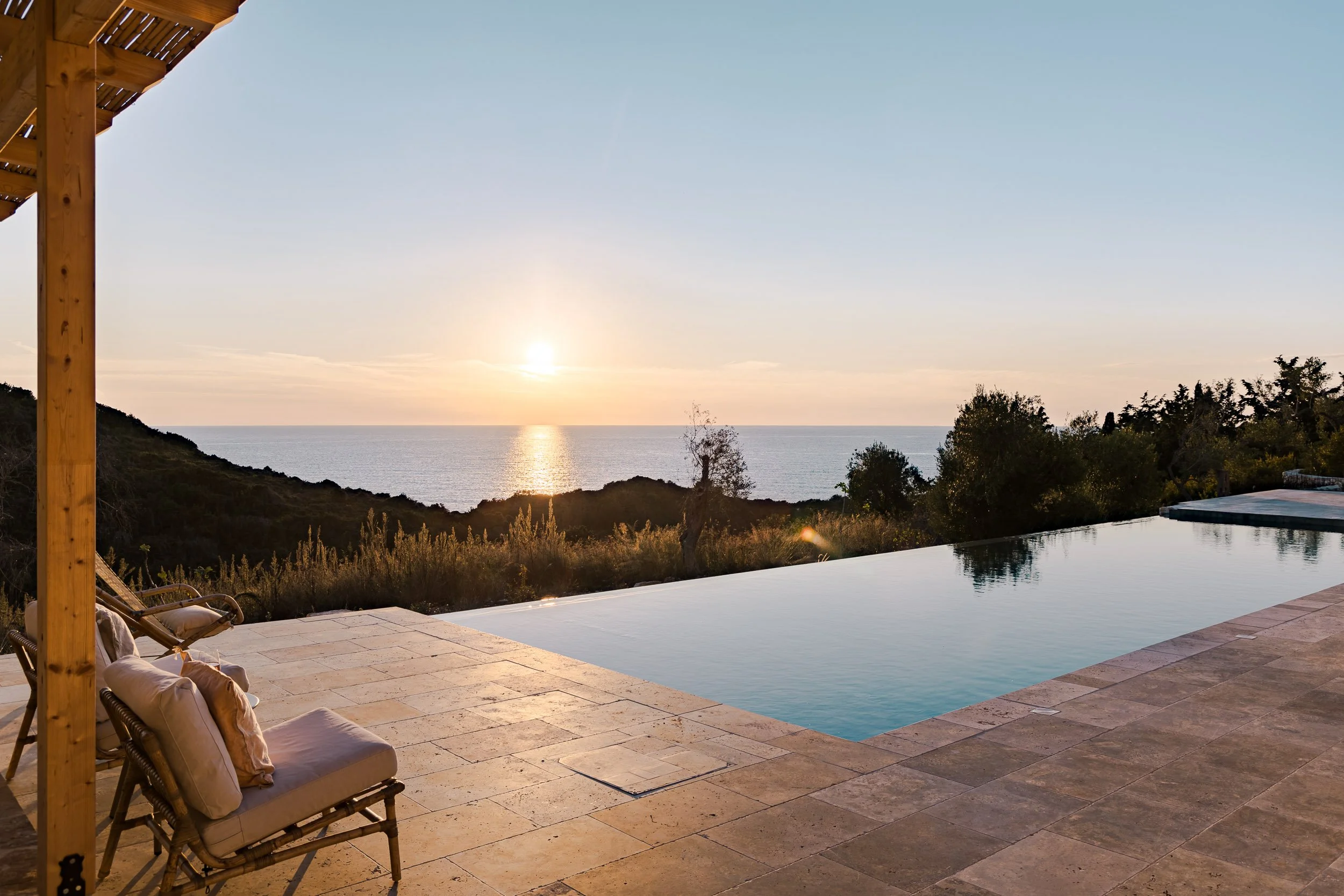 View of an infinity pool overlooking the ocean at sunset, with a hillside and trees in the background, and wicker chairs with cushions on a stone patio.