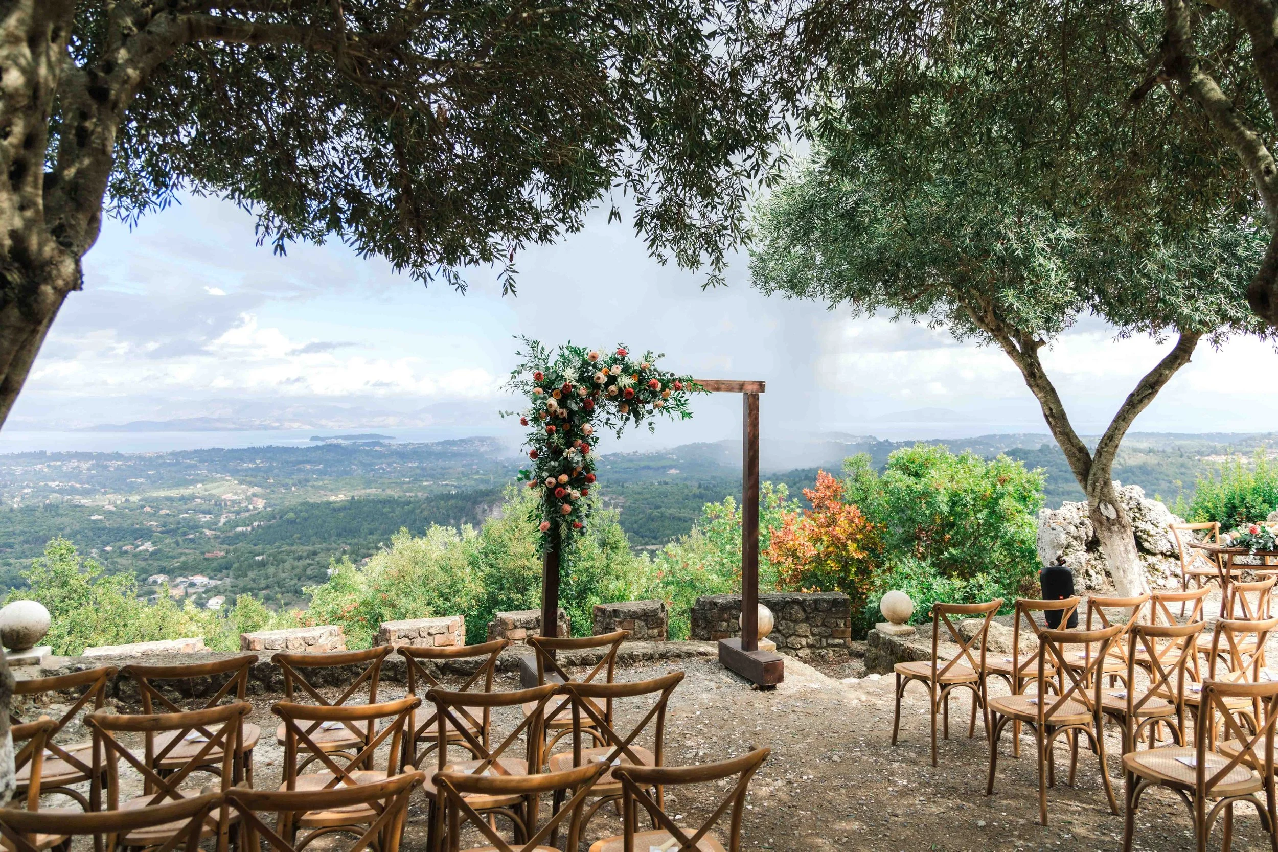 Outdoor wedding ceremony setup on a hill with wooden chairs, a floral arch, and scenic view of a valley and water in the distance, shaded by trees.