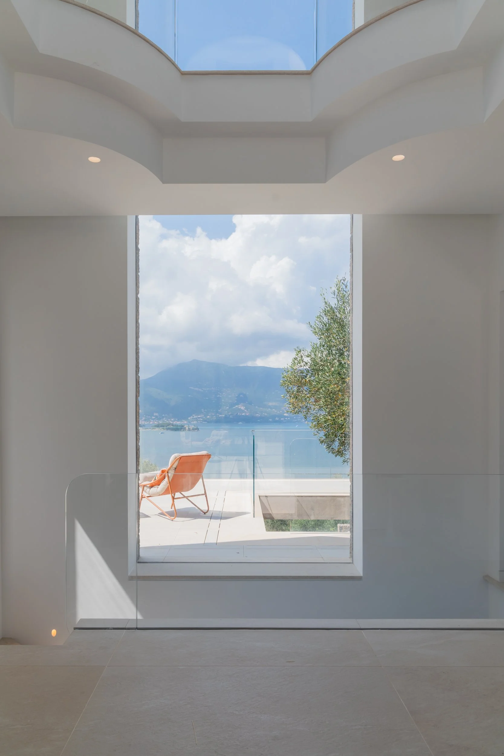 Modern interior with large window overlooking lake, mountains, and sky, with an outdoor lounge chair on patio
