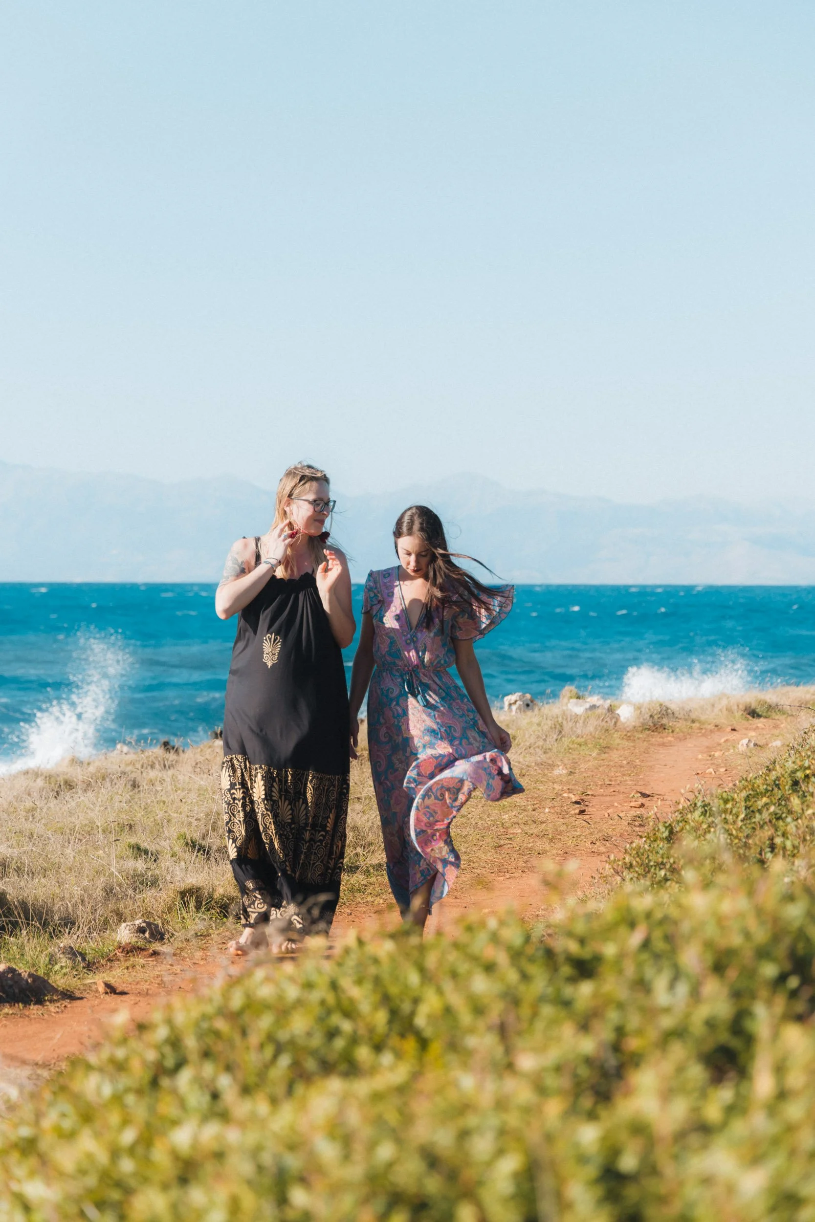 Two women walking along a dirt path near the ocean, with waves crashing in the background and hills in the distance.