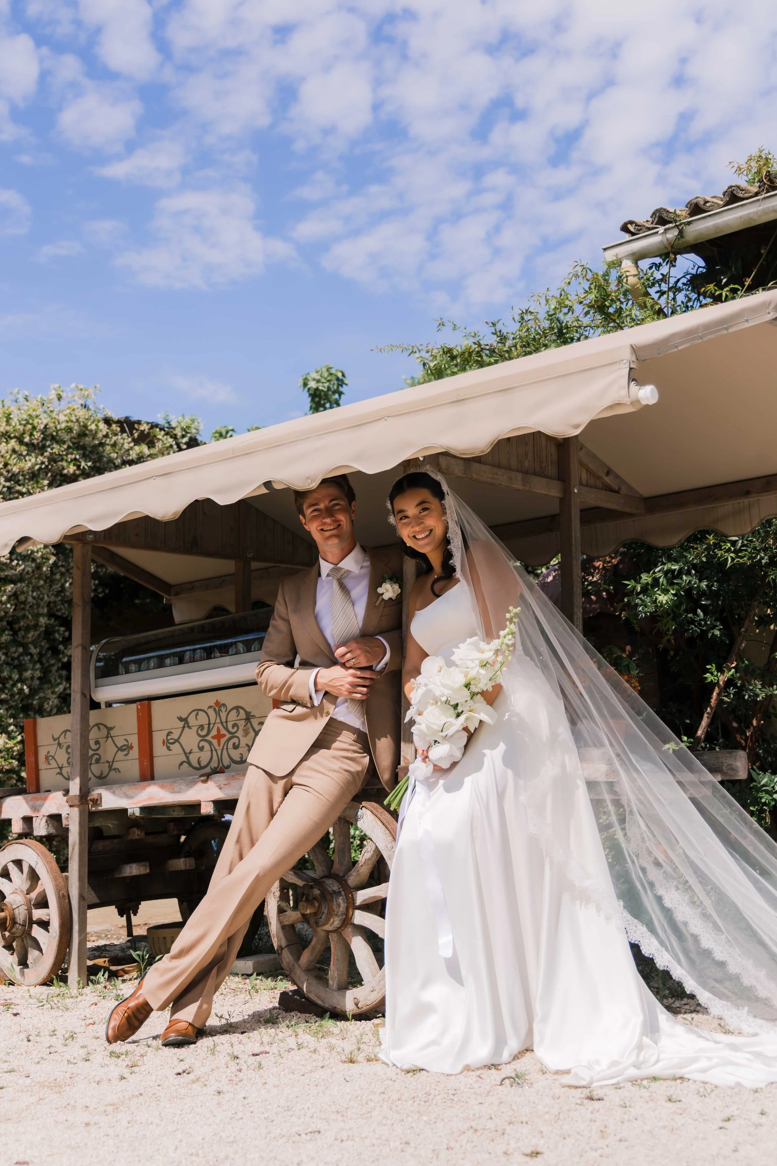 A newlywed couple in wedding attire stands outdoors under a tent, smiling at the camera with a bright sky and trees in the background.