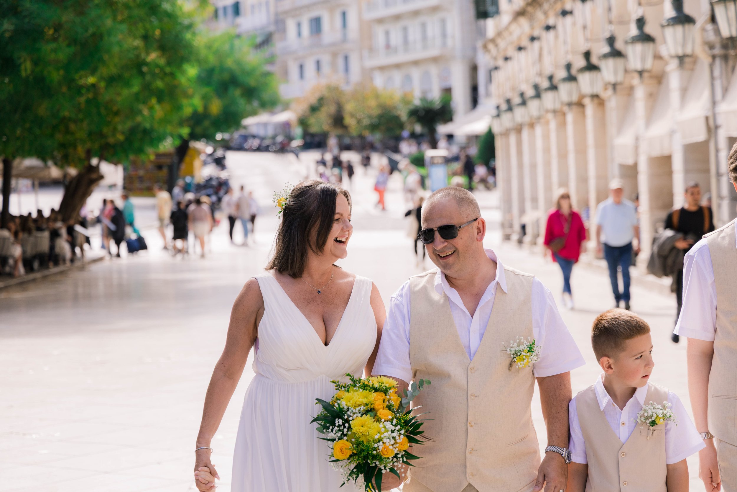 A group of people, including a woman in a white dress holding a bouquet and a man in sunglasses, walking on a city street during daytime. The woman and man are smiling and appear to be celebrating, possibly at a wedding, with other people and buildin