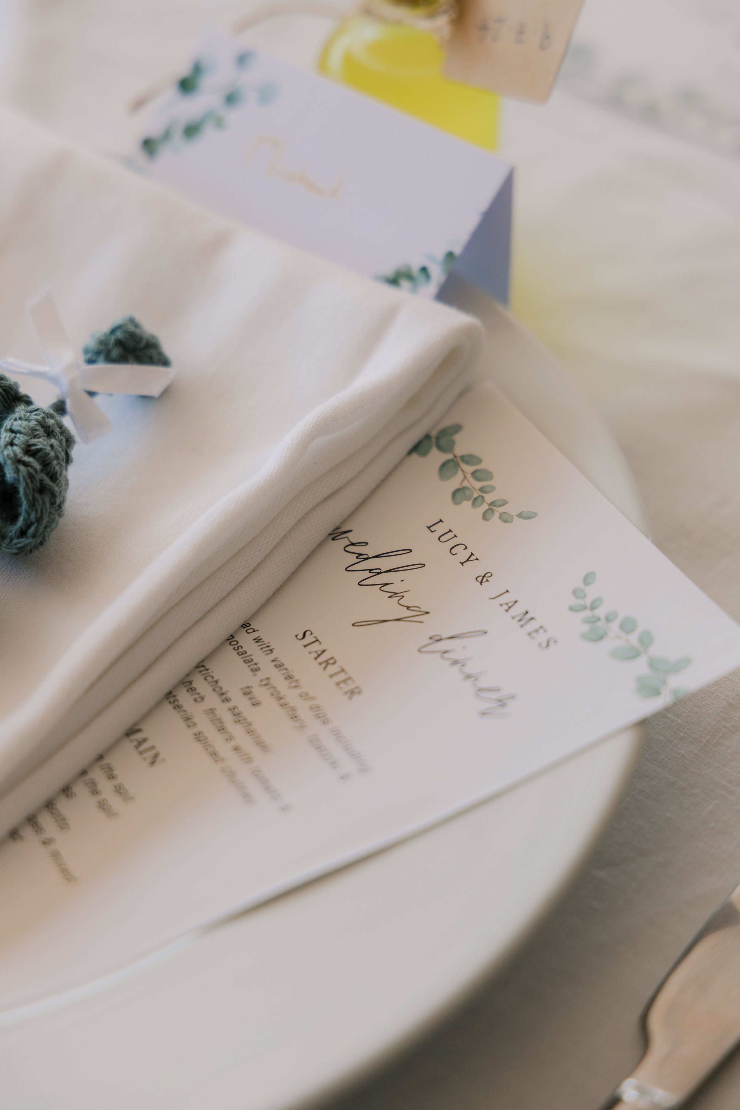 Wedding invitation card reading 'Lucy & James, Wedding Dinner, Starter, Main' decorated with green foliage designs, placed on a white table along with a folded white napkin and a white cloth.