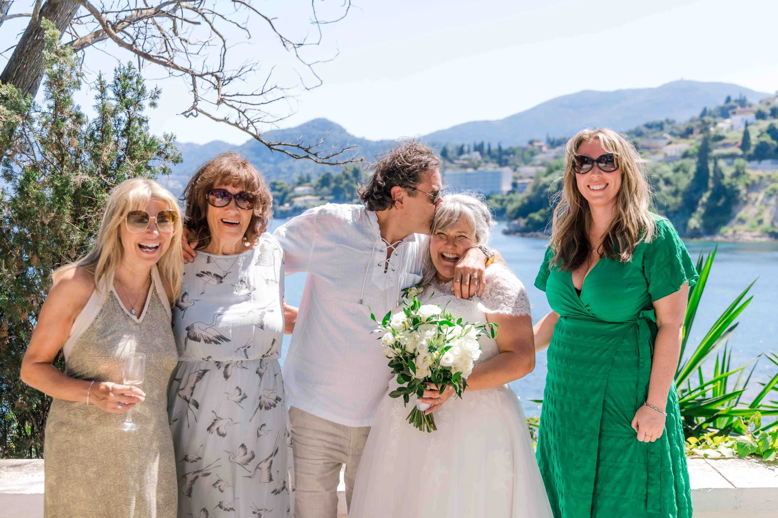 A group of six people celebrating a wedding outdoors next to a body of water with hills in the background. The bride is holding a bouquet of white flowers, and a man is kissing her on the head. The others are smiling and wearing summer dresses and su
