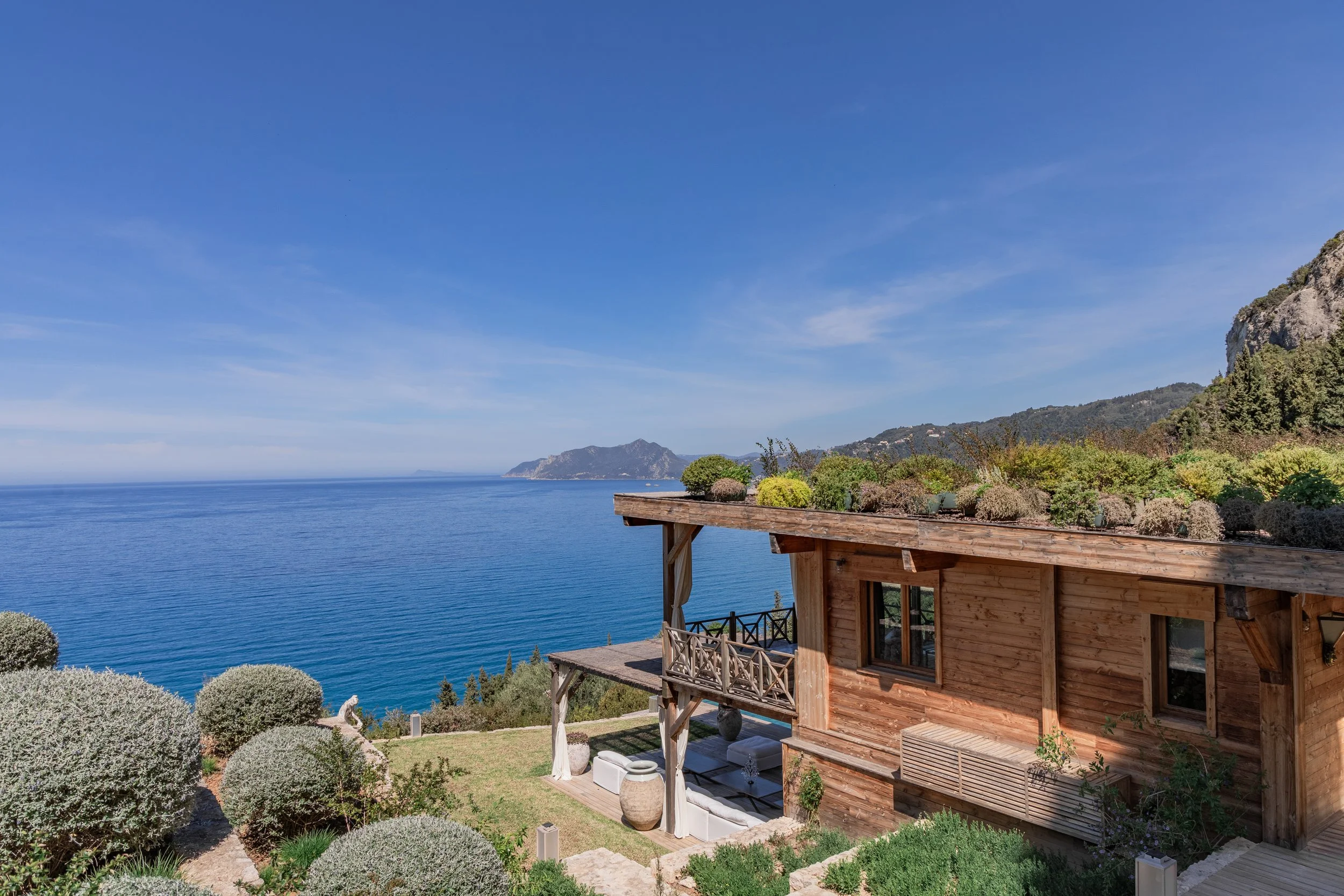 A wooden house overlooking the ocean, with a balcony and lush garden in the foreground, mountains in the distance under a blue sky.