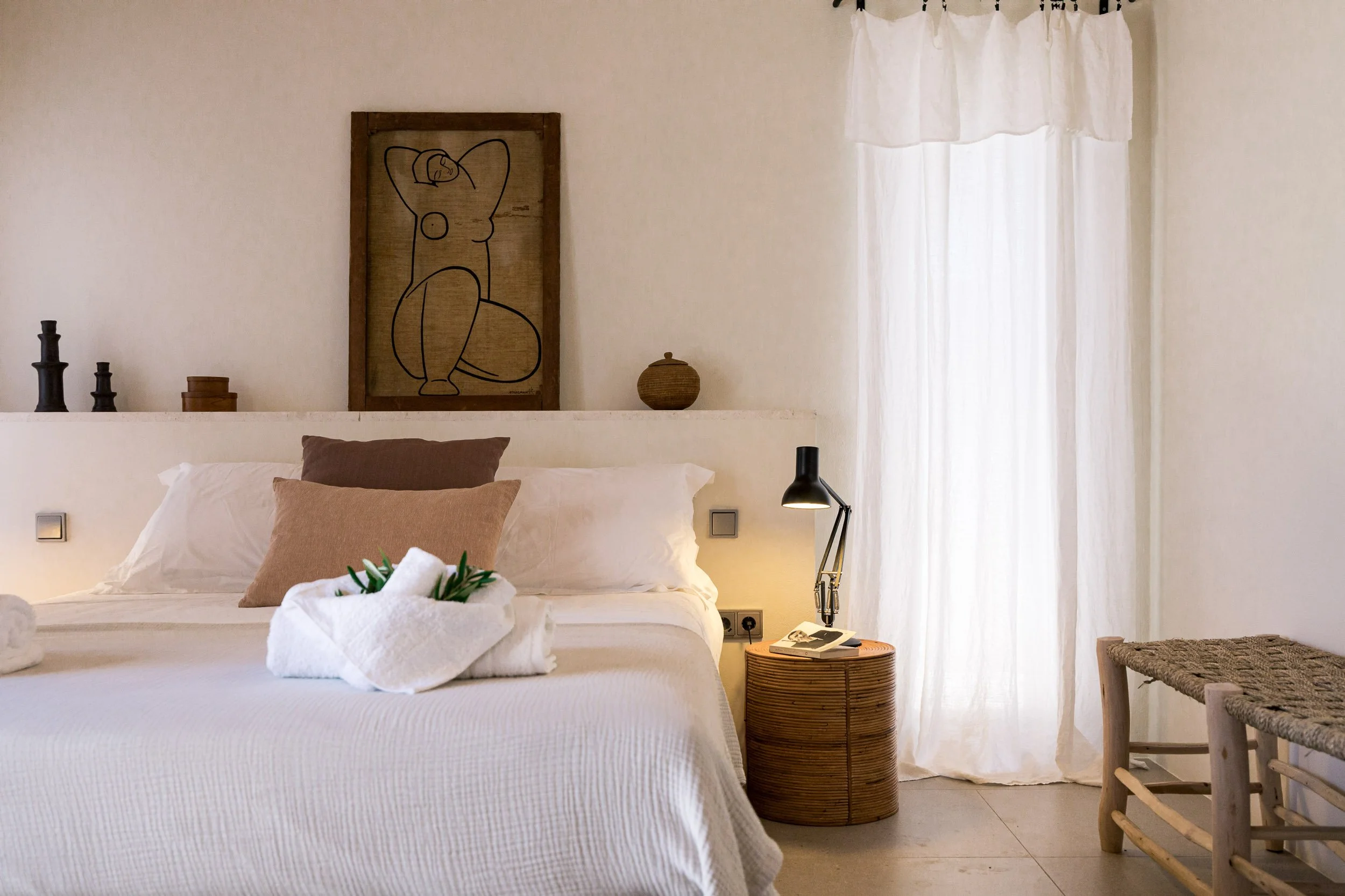 A bedroom with a white bed, brown and beige pillows, a towel shaped like a swan with green leaves on it, a nightstand with a black desk lamp and phone, sheer white curtains, a wooden bench, and decorative items including a framed line art of a nude f
