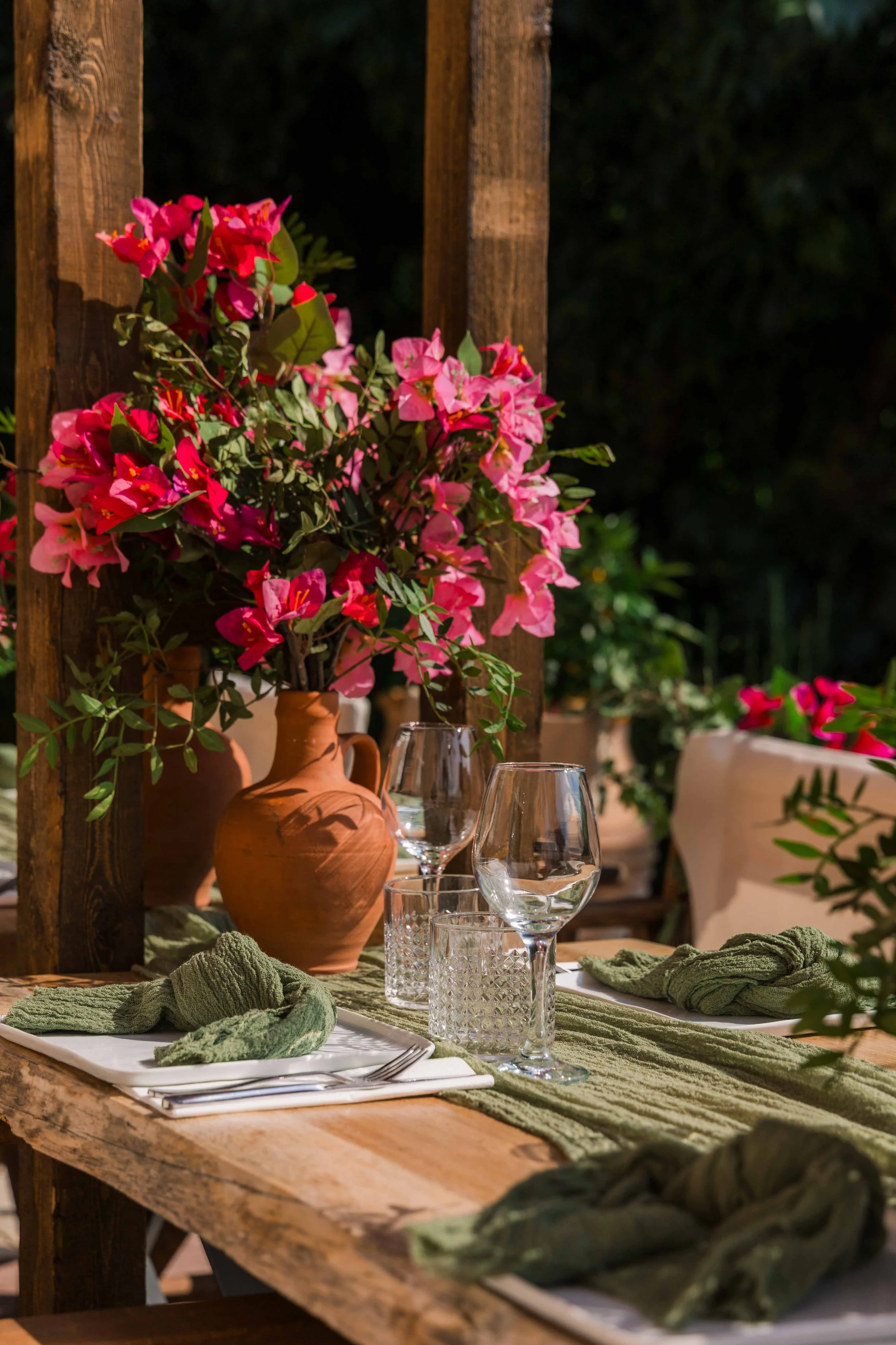 A rustic outdoor dining table with a large terracotta vase filled with pink and red bougainvillaea flowers, set with green napkins, glassware, and a green table runner, with lush greenery in the background.