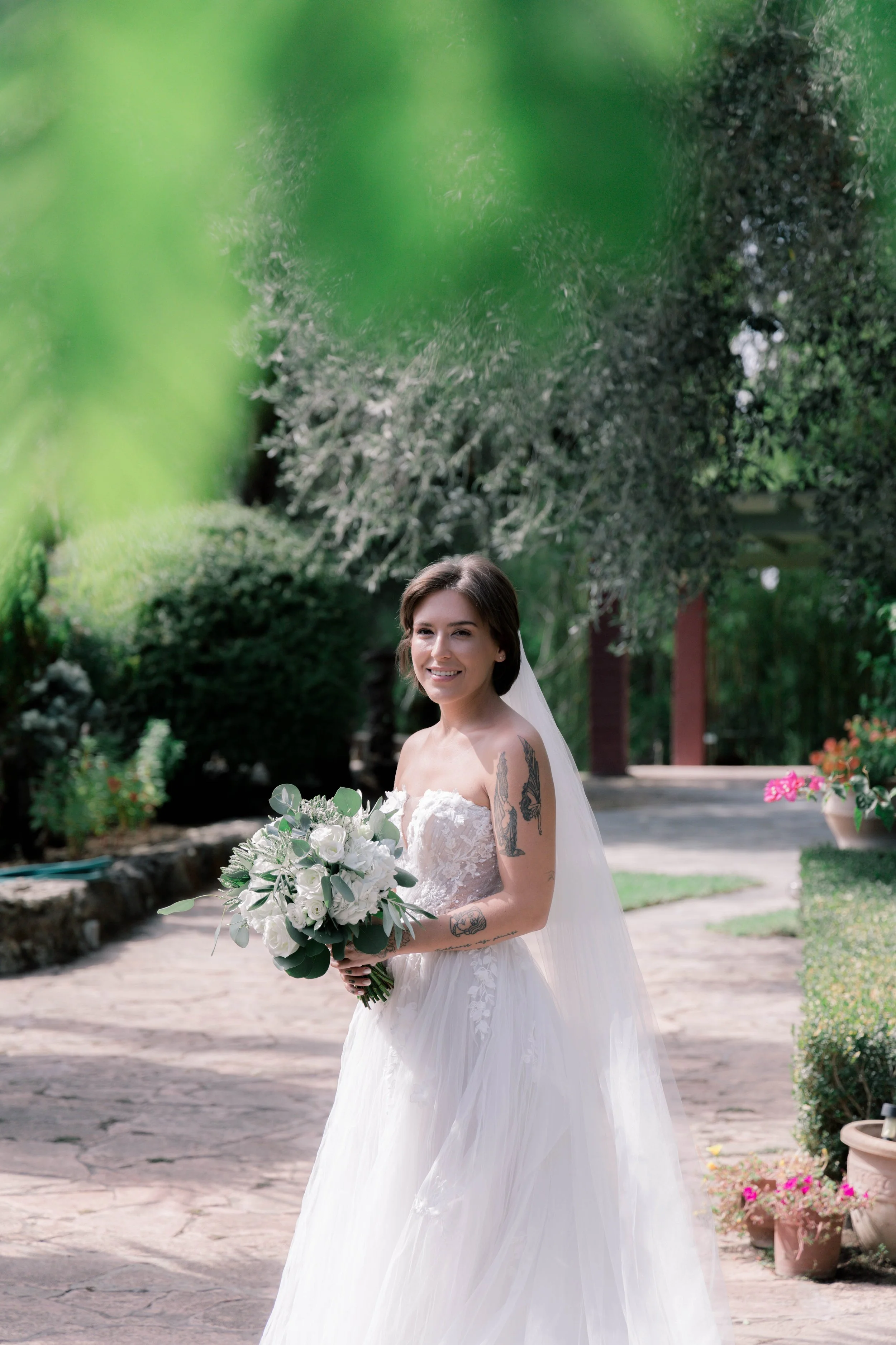 A bride in a strapless white wedding gown holding a bouquet of white flowers and greenery, smiling outdoors in a garden setting with trees, bushes, and flower pots.