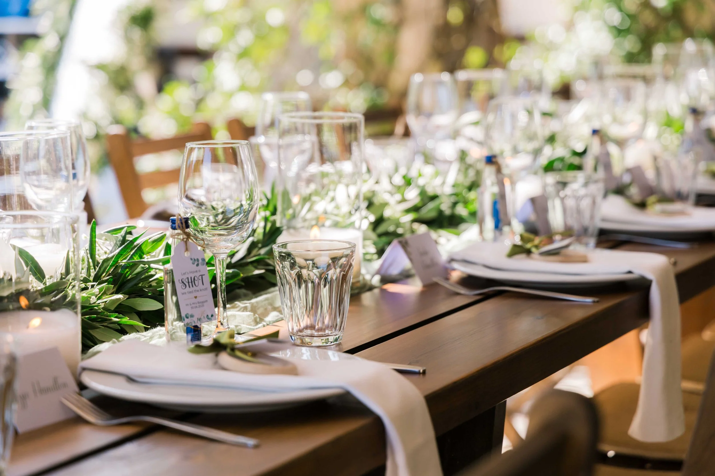 A decorated outdoor dining table set for a special event, featuring glassware, napkins, cutlery, candles, and a lush green floral centerpiece.
