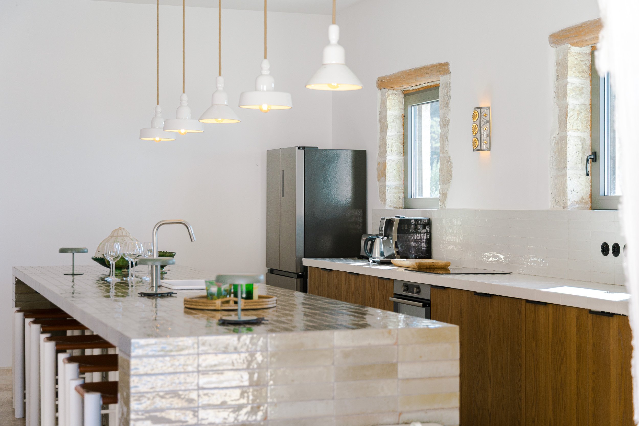 Modern kitchen with white tiled island, four wine glasses, and minimalist hanging pendant lights, featuring stainless steel refrigerator, countertop appliances, and two small windows with stone framing.