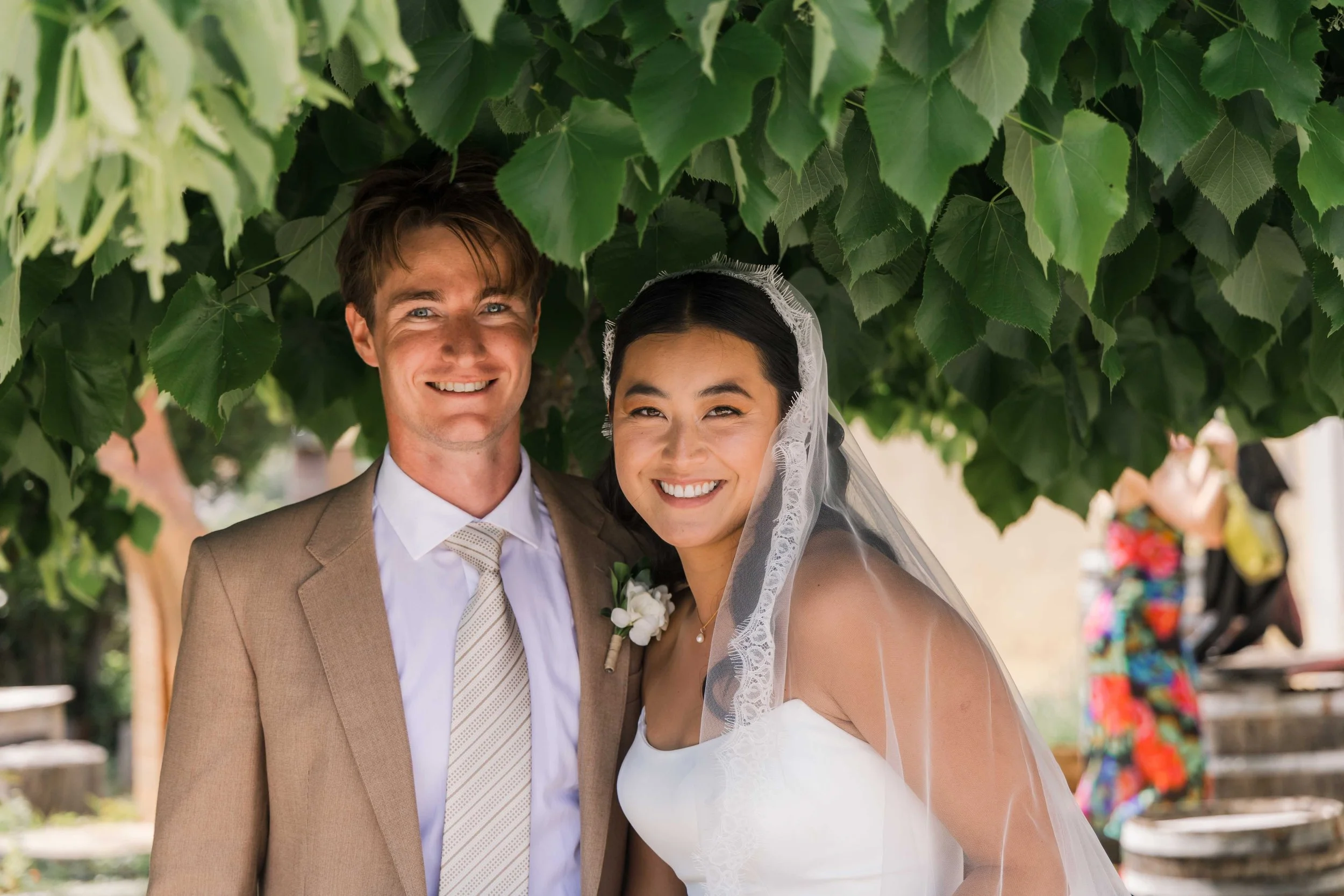 A newlywed couple smiling under a leafy canopy, with the bride wearing a white wedding dress and veil, and the groom in a tan suit and white shirt.