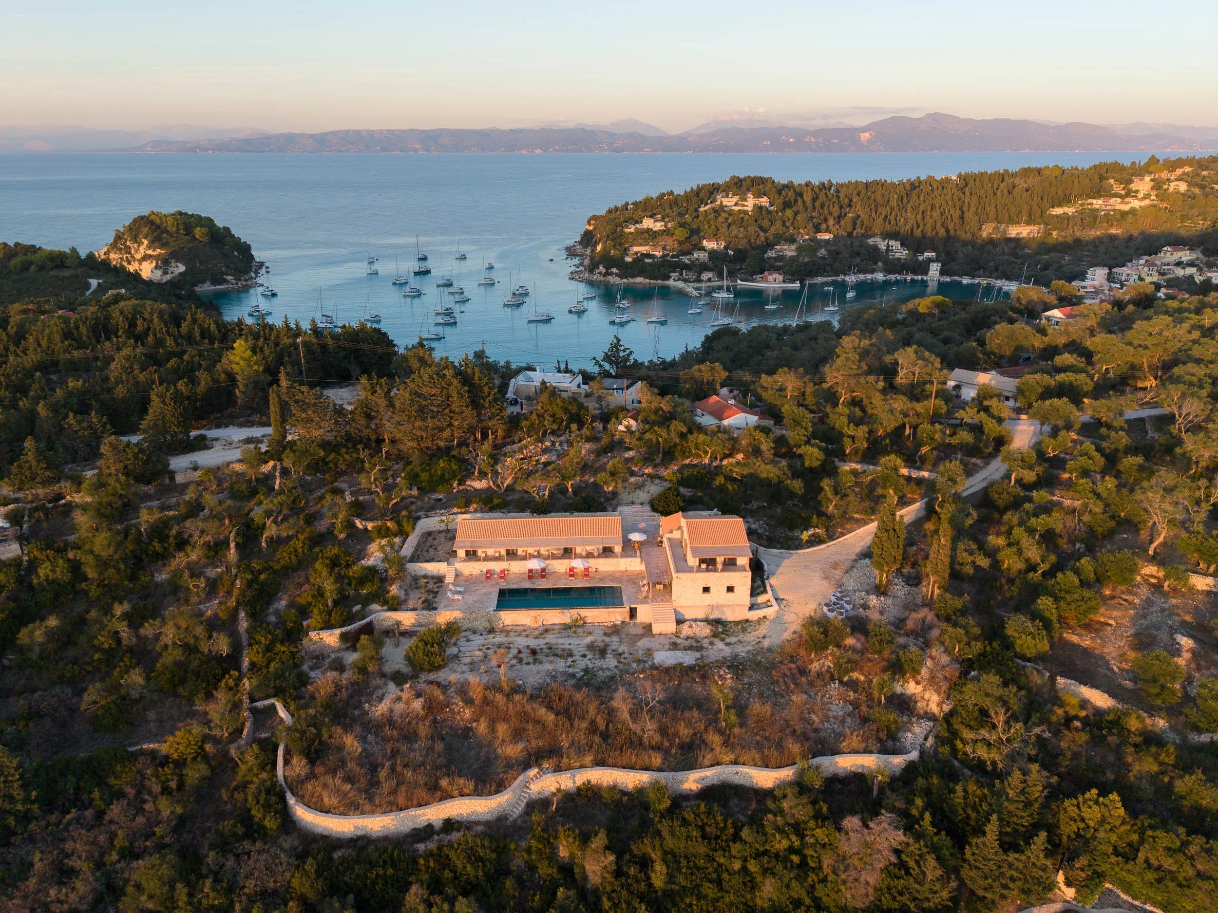 Aerial view of a coastal area with a private house, a swimming pool, and a winding boundary wall surrounded by trees. In the background, there is a bay with sailboats and a distant mountainous shoreline.