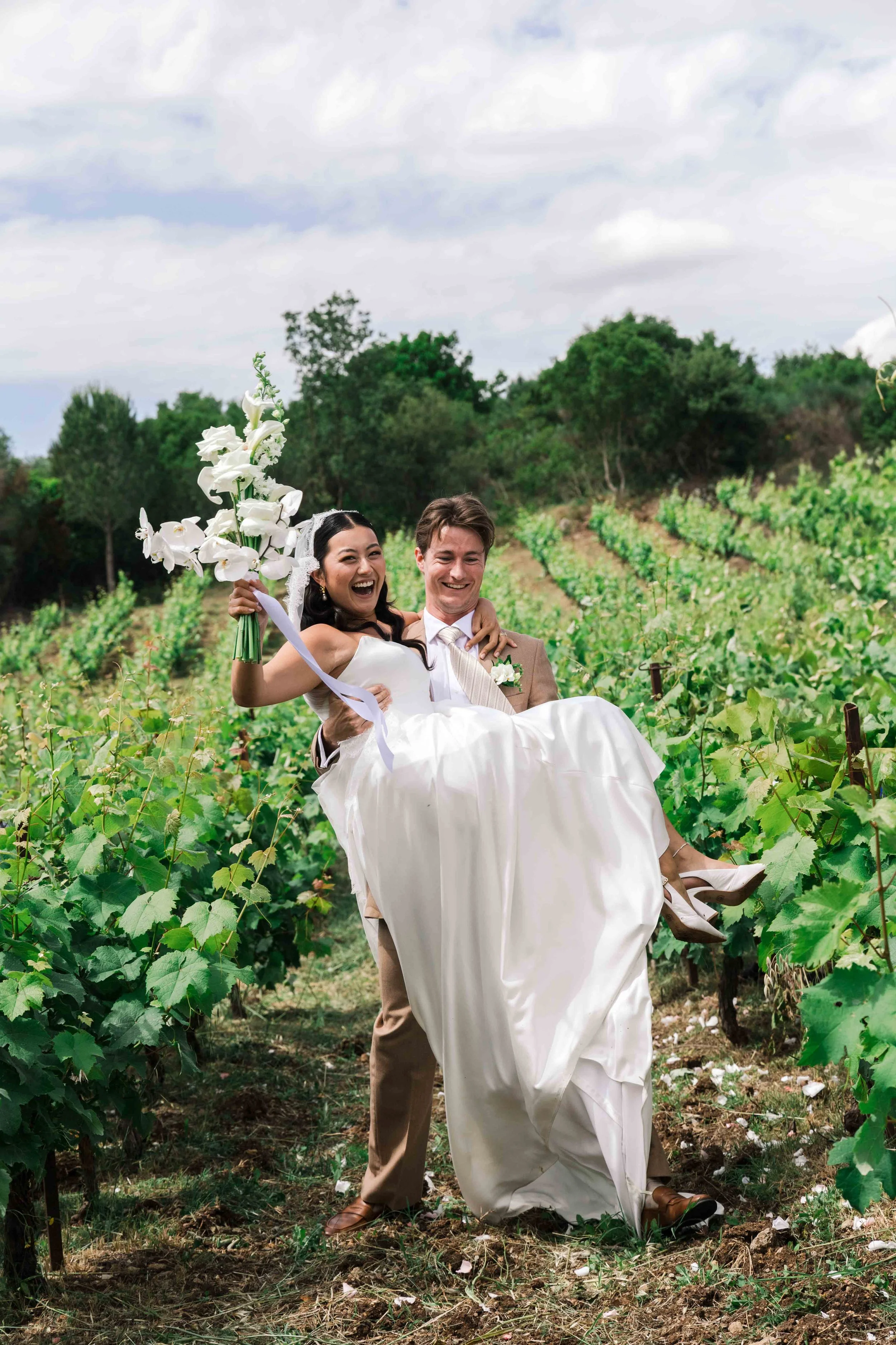 A newlywed couple in wedding attire, with the groom carrying the bride, smiling happily in a vineyard setting.