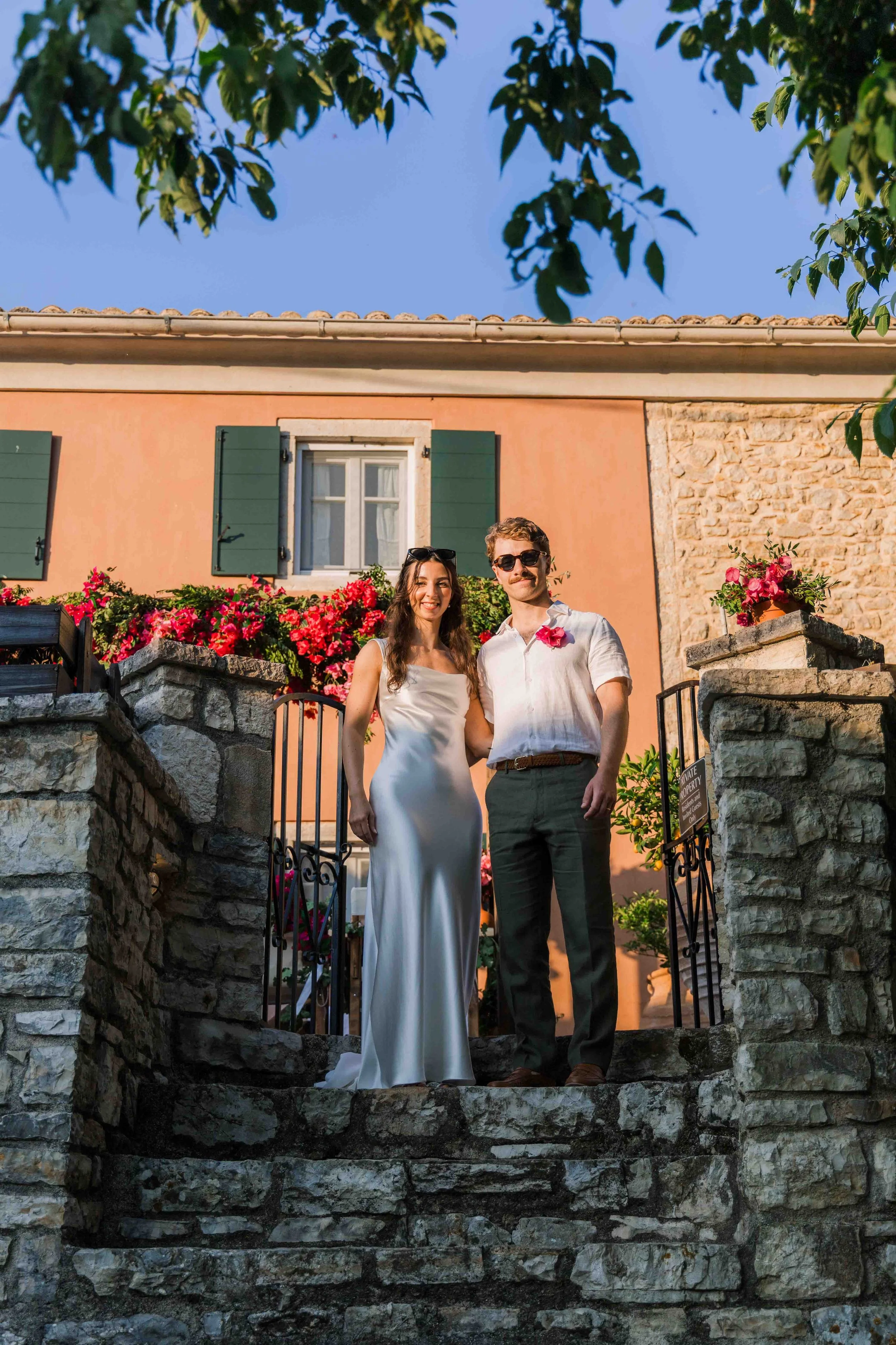 A couple standing on stone steps in front of a pink house with green shutters and blooming pink flowers. The woman is wearing a white satin dress, and the man is in a white shirt and black pants, both wearing sunglasses. The setting suggests a romant