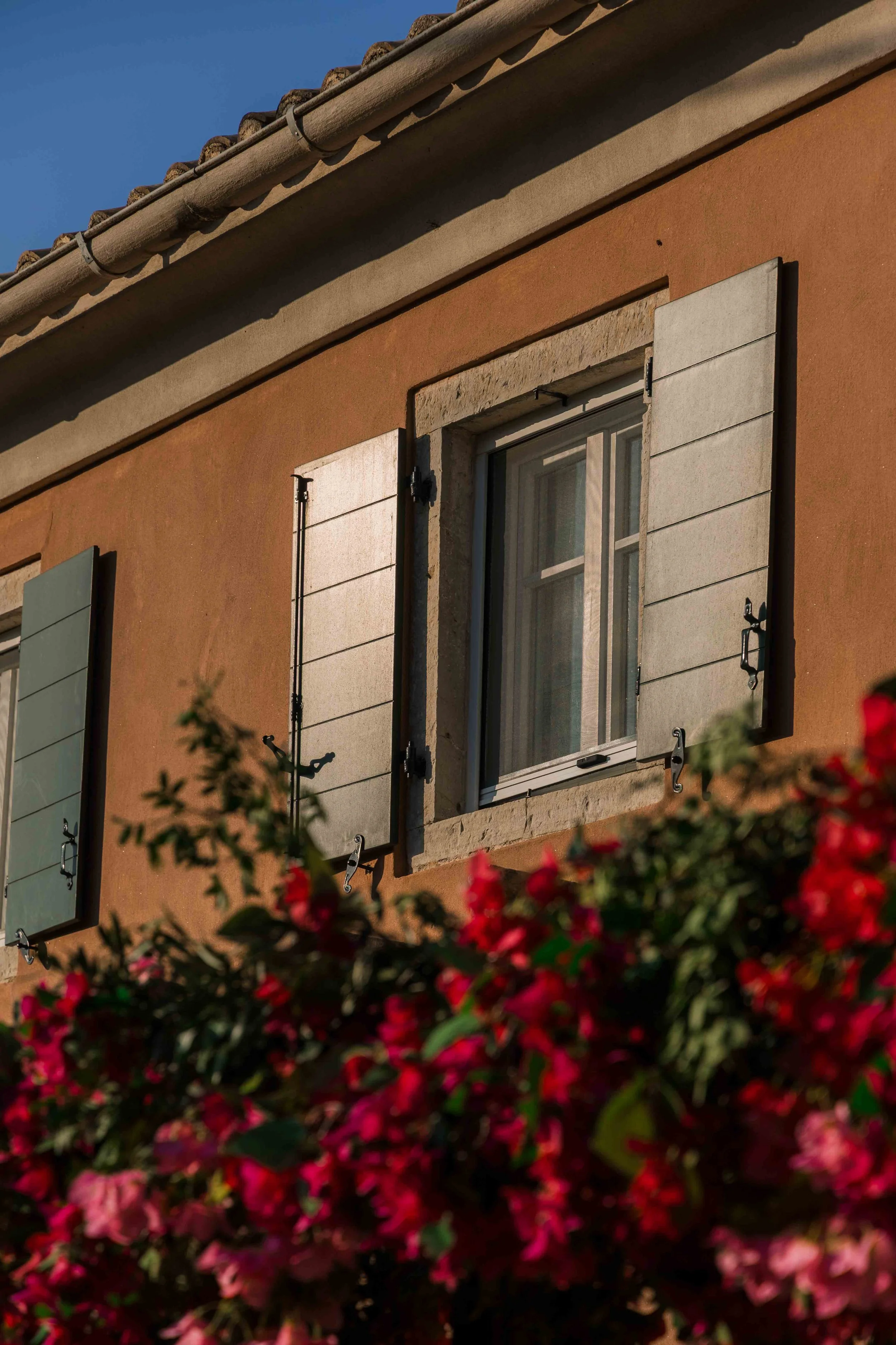 A close-up of an orange stucco house wall with two windows, each with open white shutters, and pink flowering bushes in the foreground. The house has a tiled roof under a clear blue sky.