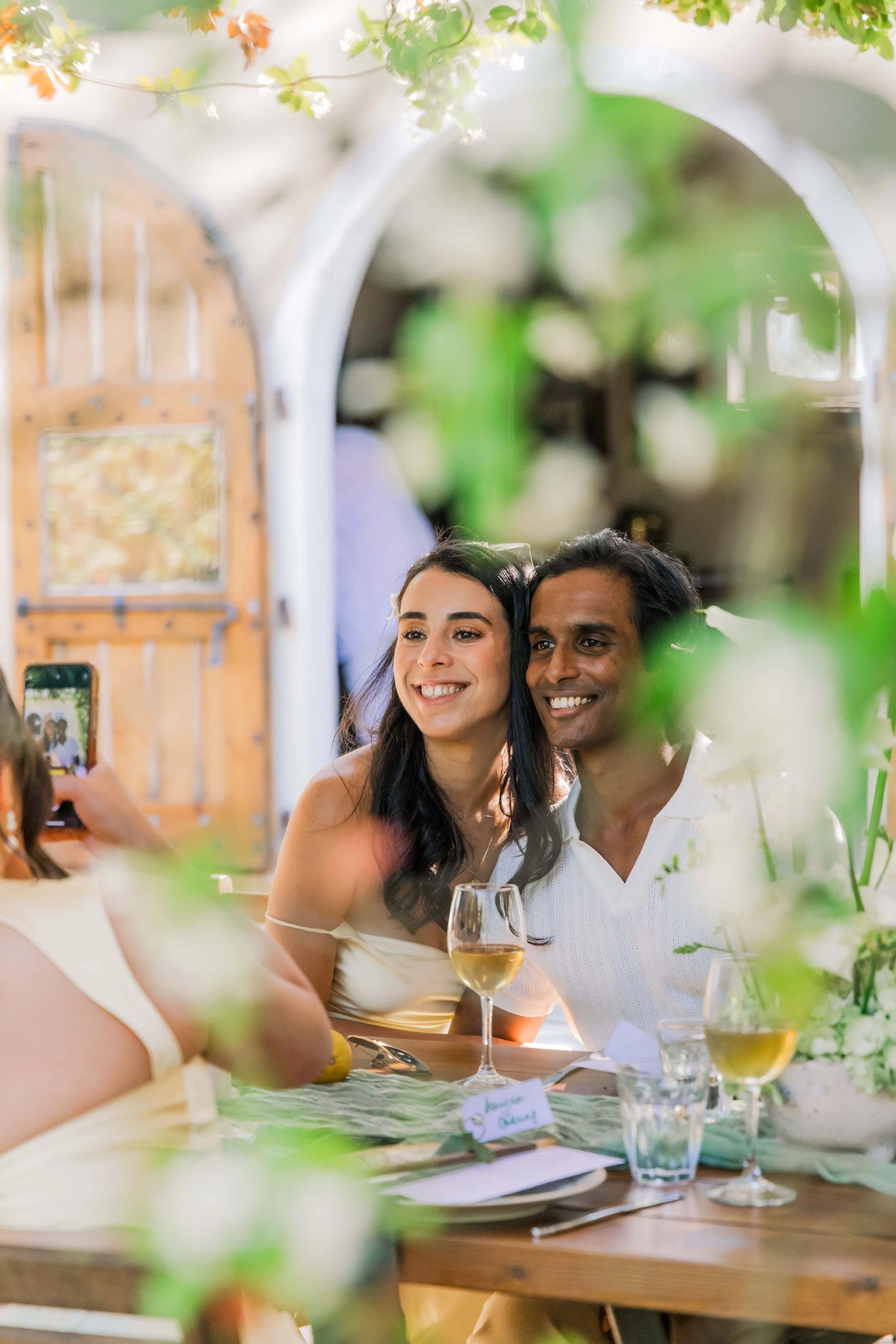 Smiling couple taking a selfie at a dinner table decorated with flowers and wine glasses.