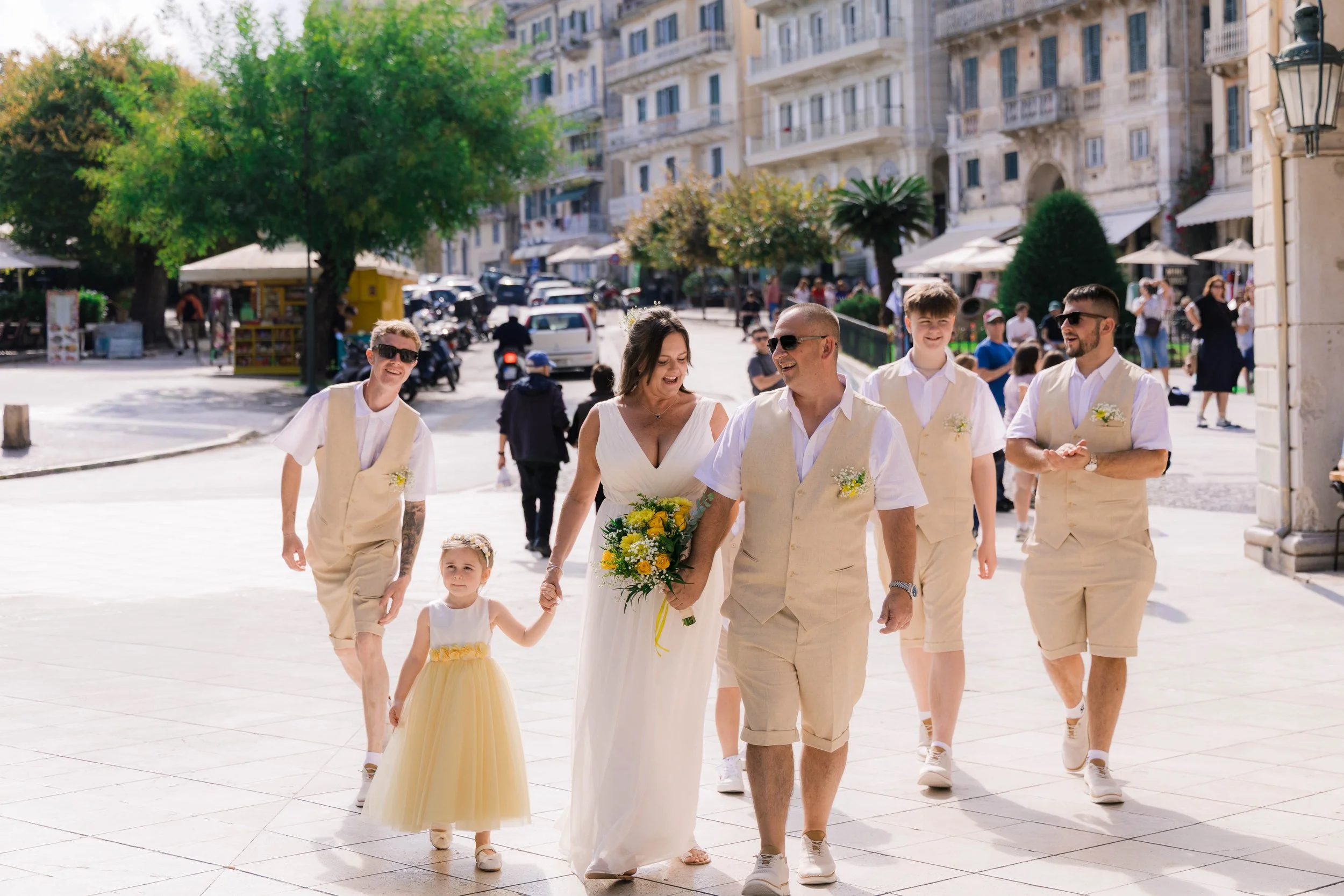 A group of people, including a bride holding a bouquet, walking outdoors on a sunny day in a city square. The group includes children and adults dressed in light-colored, casual wedding attire, smiling and enjoying the moment.