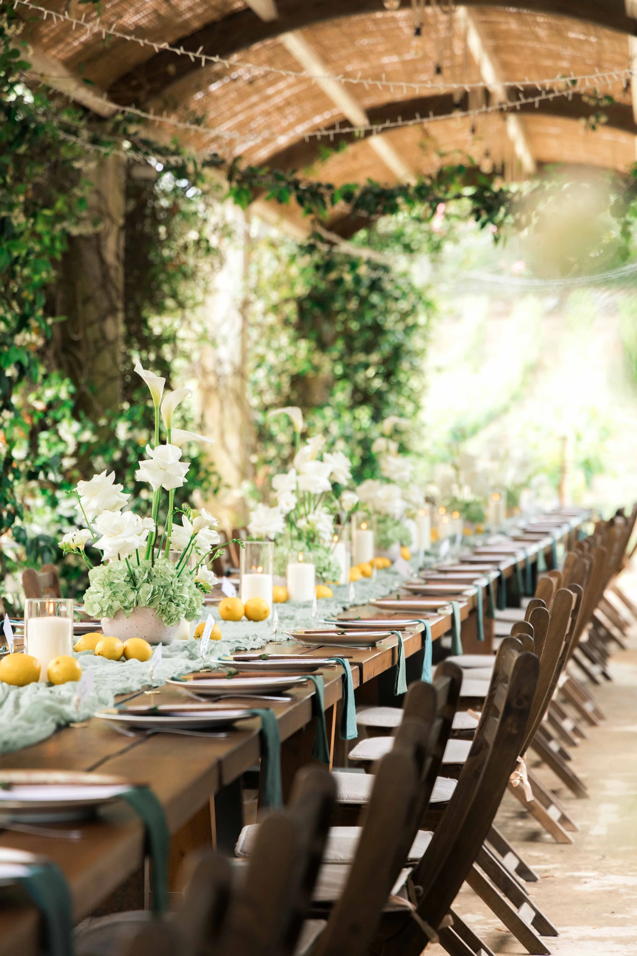 A long outdoor dining table decorated with white flowers, candles, and lemons under a thatched roof with string lights, surrounded by greenery.