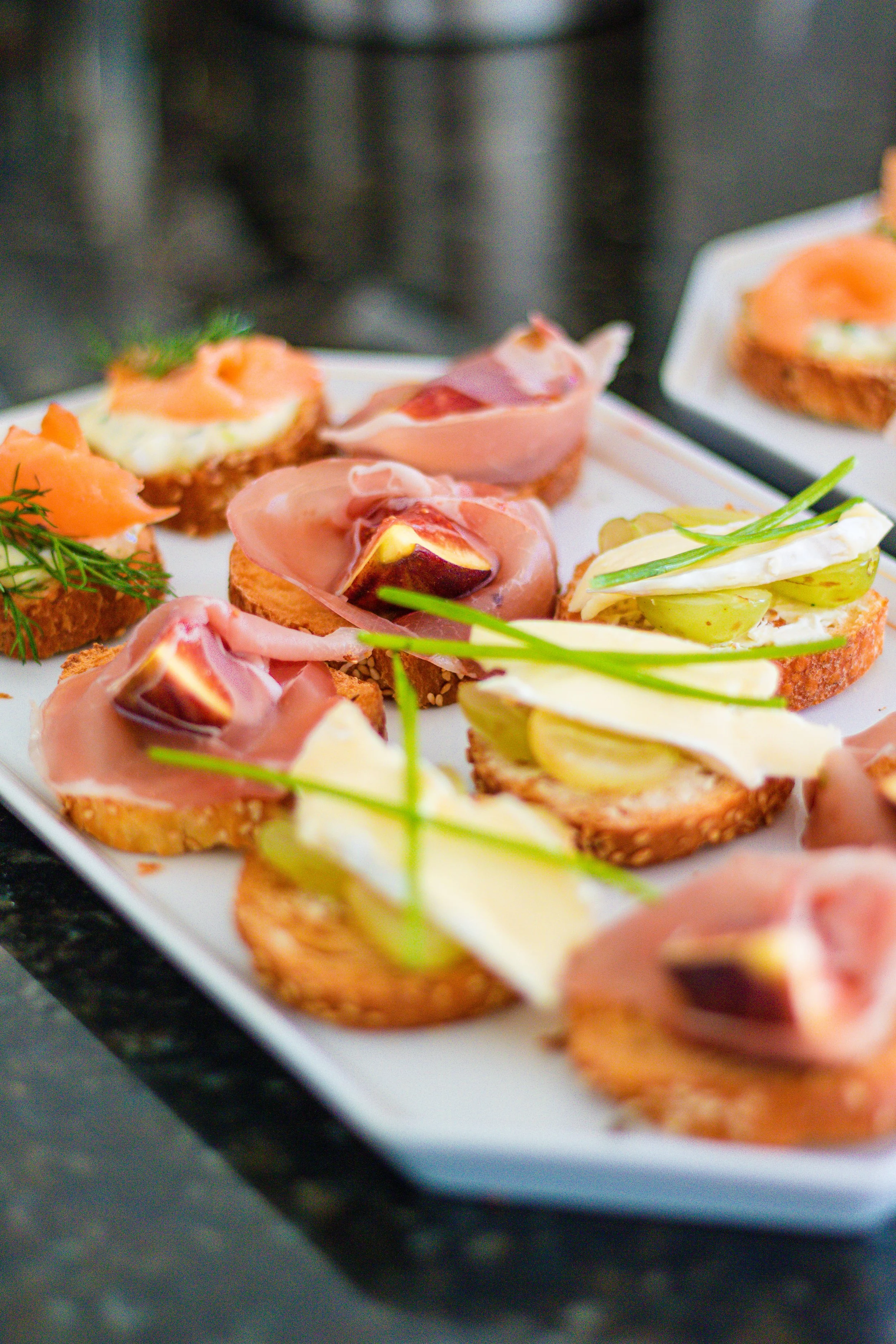 Assorted open-faced appetizers on a white platter, topped with smoked salmon, prosciutto, cheese, grapes, and herbs.