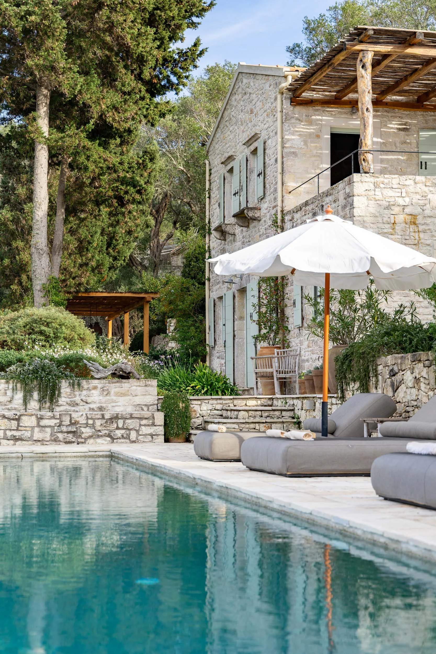 Luxury house with stone walls, outdoor pool, lounging chairs, and a white patio umbrella surrounded by greenery.