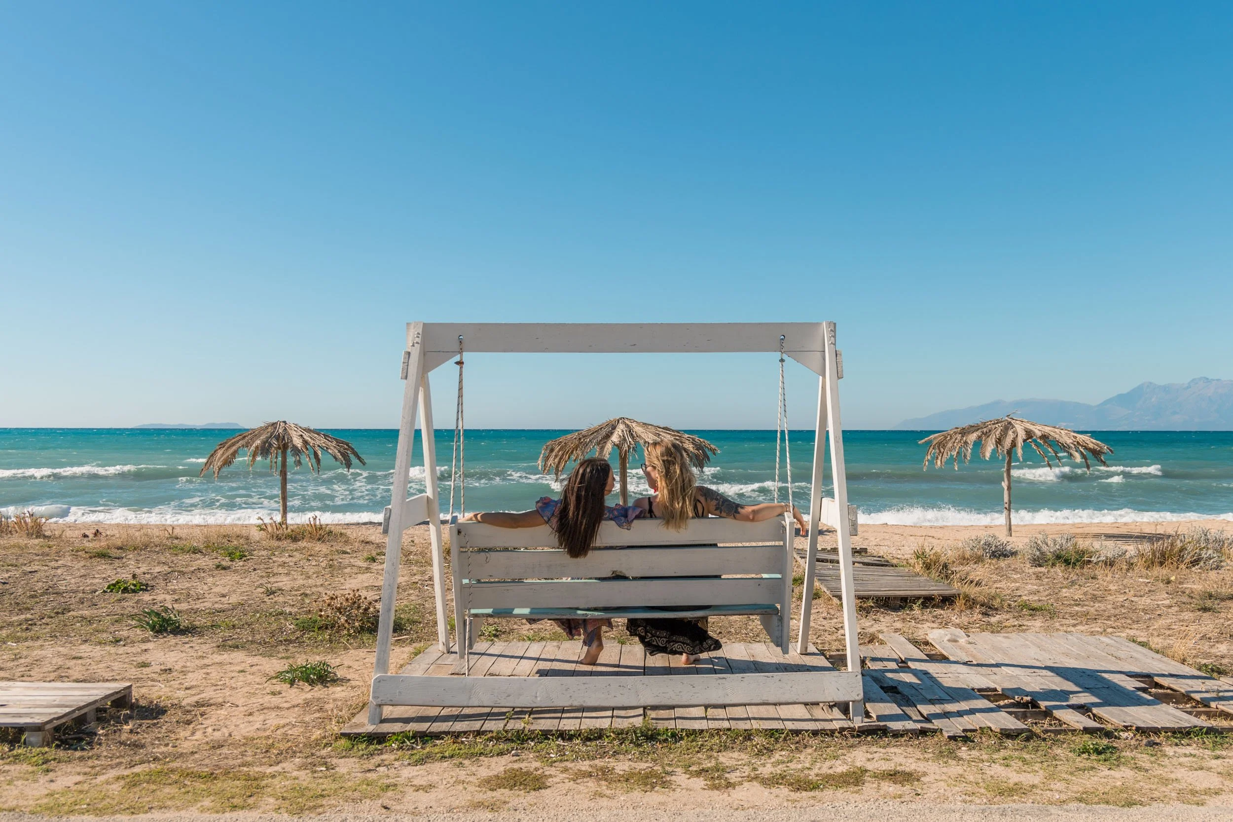 Two women sitting on a white porch swing on the beach, looking at each other and smiling, with straw beach umbrellas and the ocean in the background.