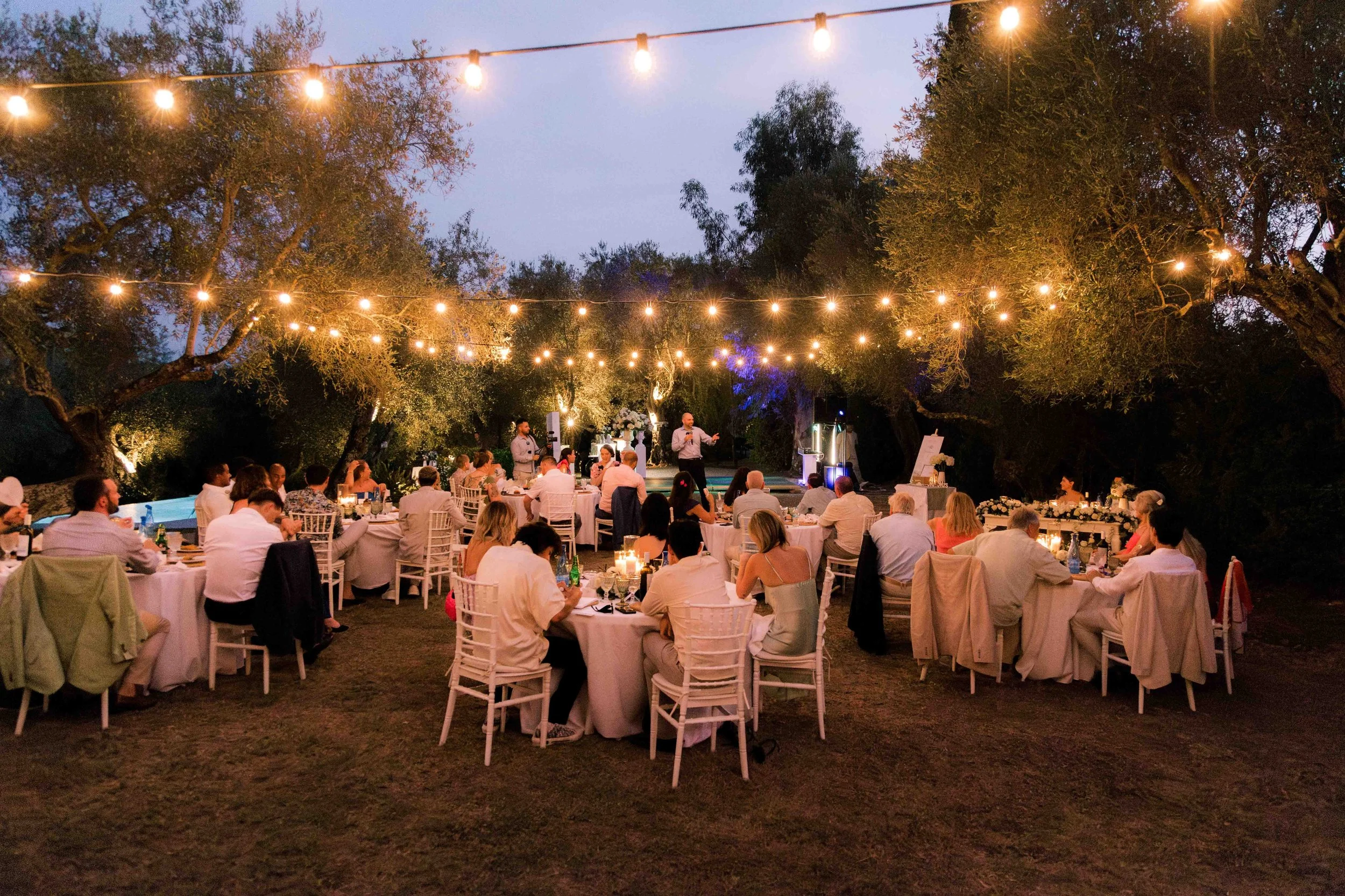 An outdoor evening celebration with round tables, white chairs, and string lights hanging overhead. People are seated at the tables, dining and socializing, with a small stage area where a speaker is addressing the crowd. Trees surround the area, cre