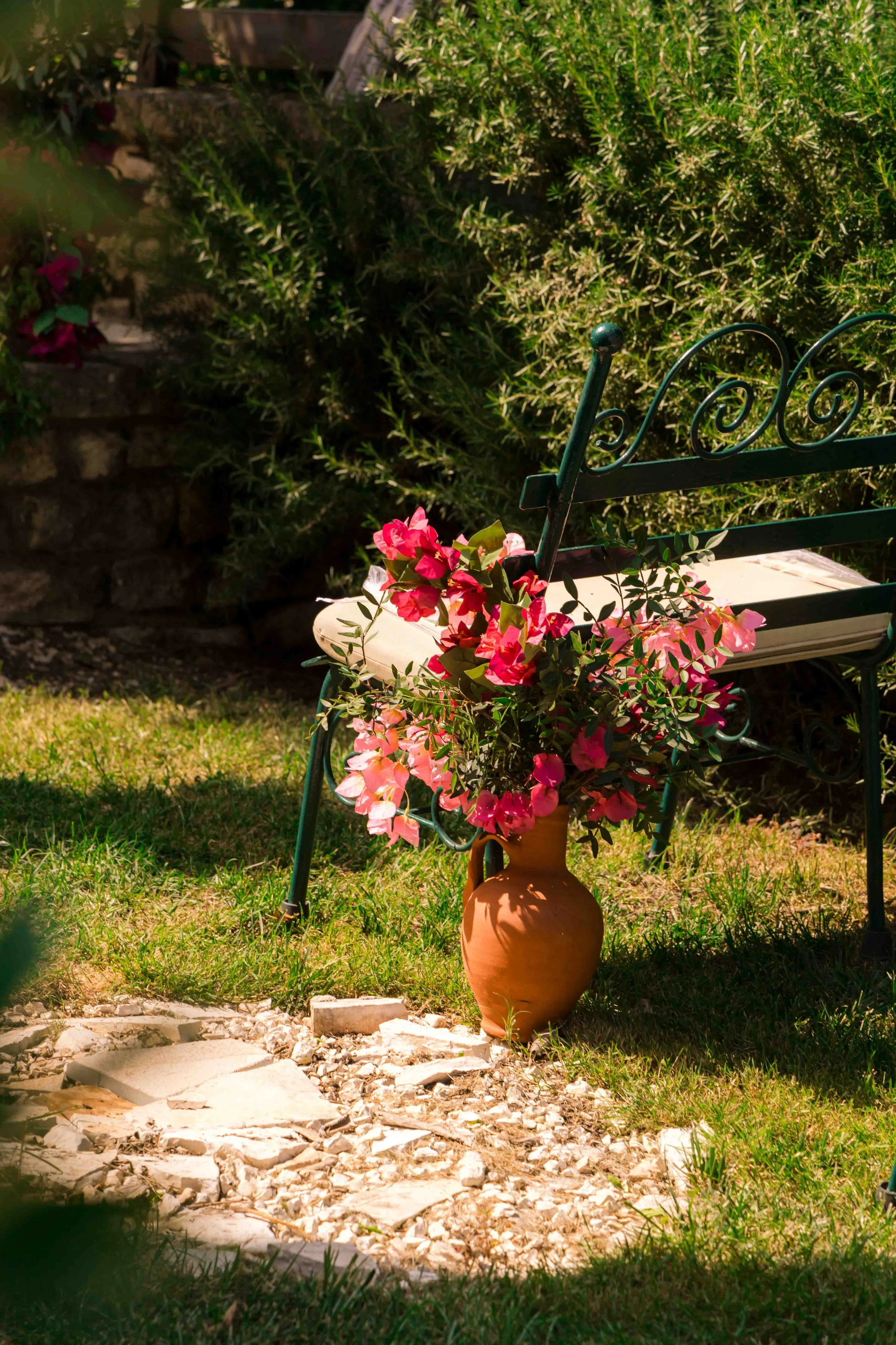 A terracotta vase filled with pink and red flowers placed on the ground next to a green garden bench surrounded by green bushes.