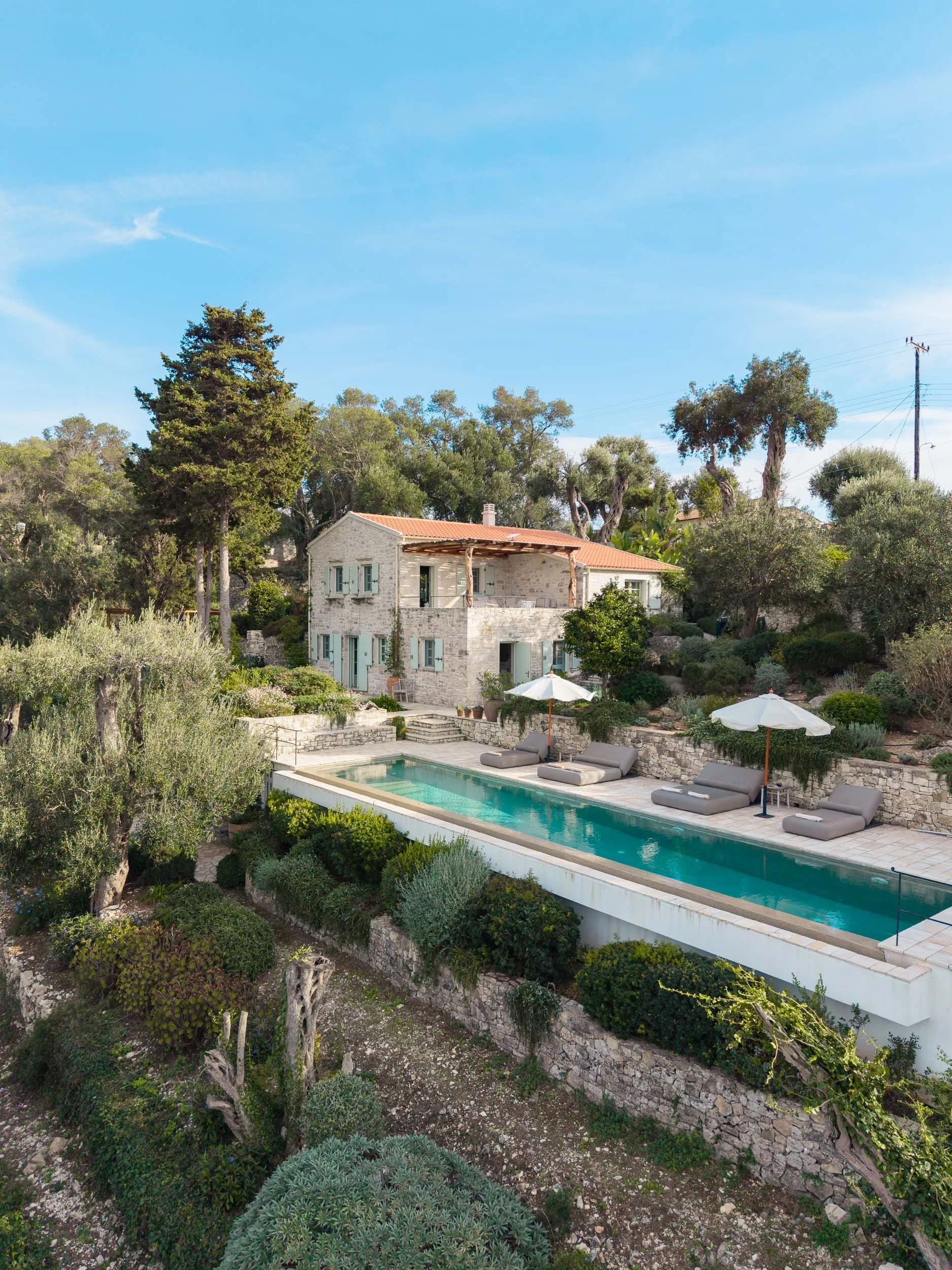 A house with a red roof, white walls, and blue shutters, surrounded by trees and garden, with a swimming pool, umbrellas, and lounge chairs outside.