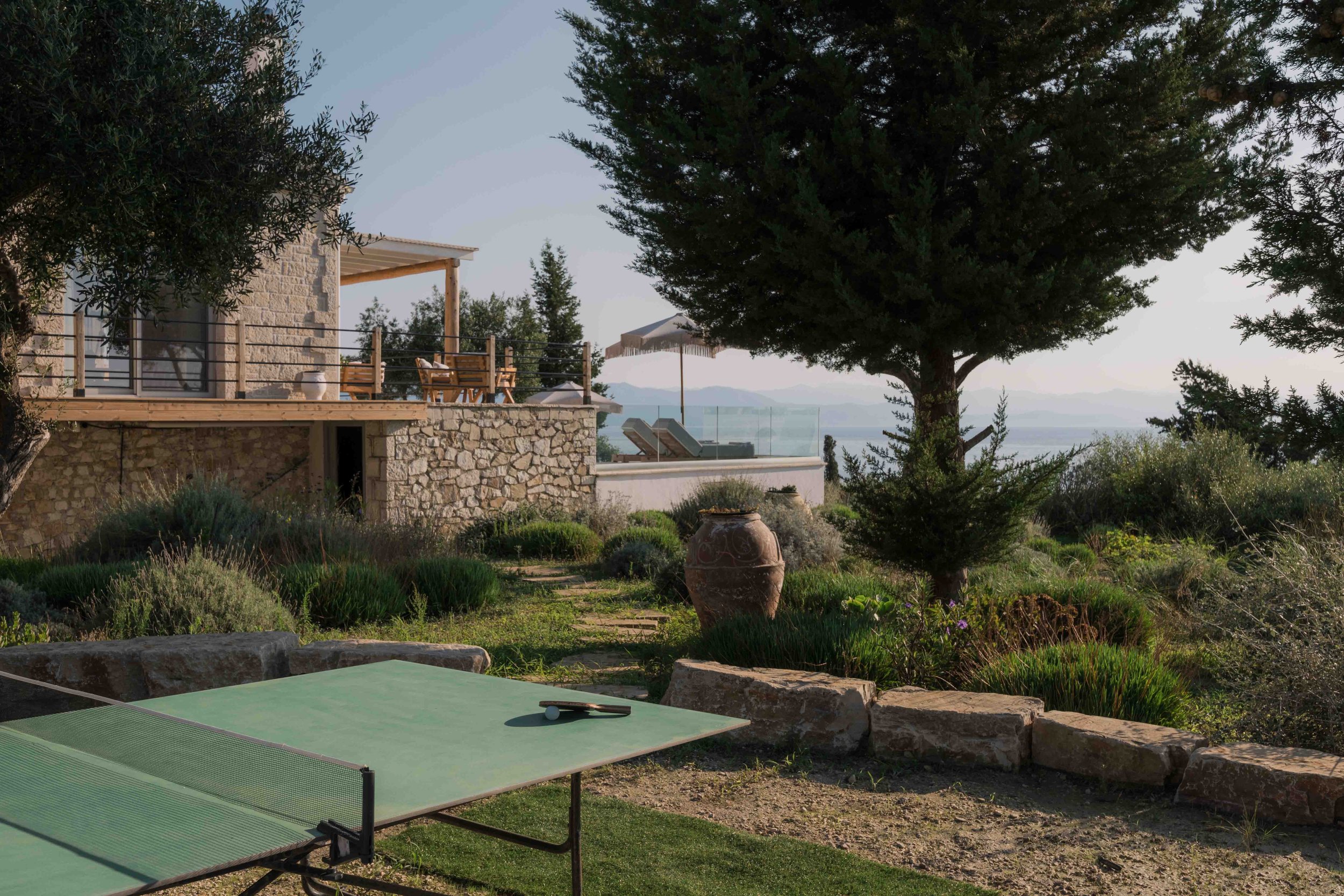 Outdoor terrace with a table tennis table, a stone house with a balcony, lush greenery, trees, and a view of distant mountains and water.